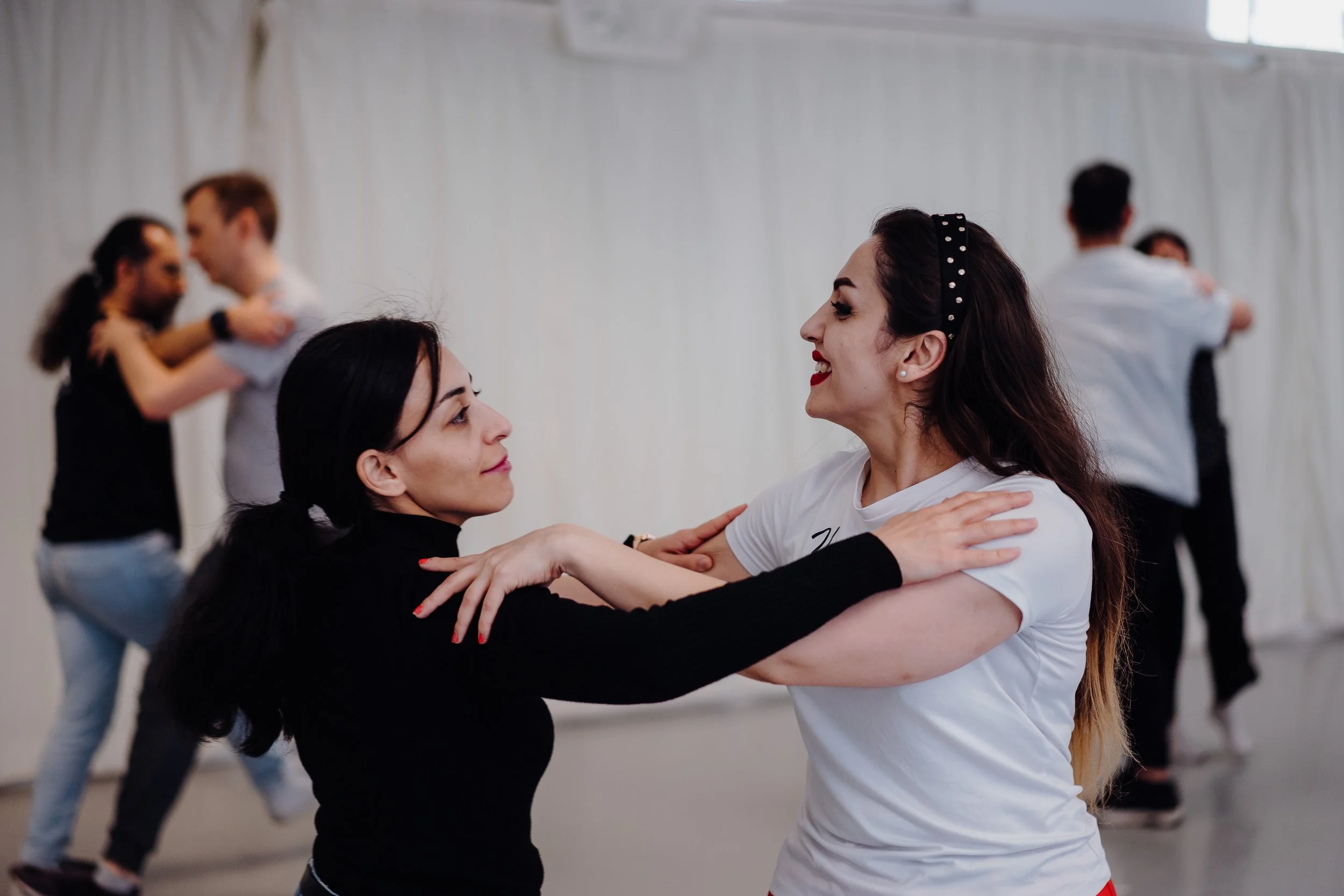 Two women dancing closely with smiles, holding each other's shoulders. Two couples in the background dancing in a brightly lit room with a white wall.