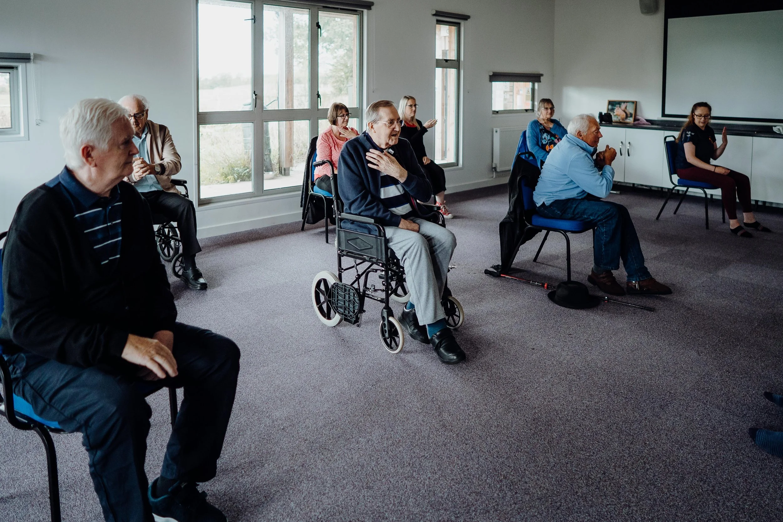 Group of elderly people in a seated circle, some in wheelchairs, participating in an activity in a community room with large windows.