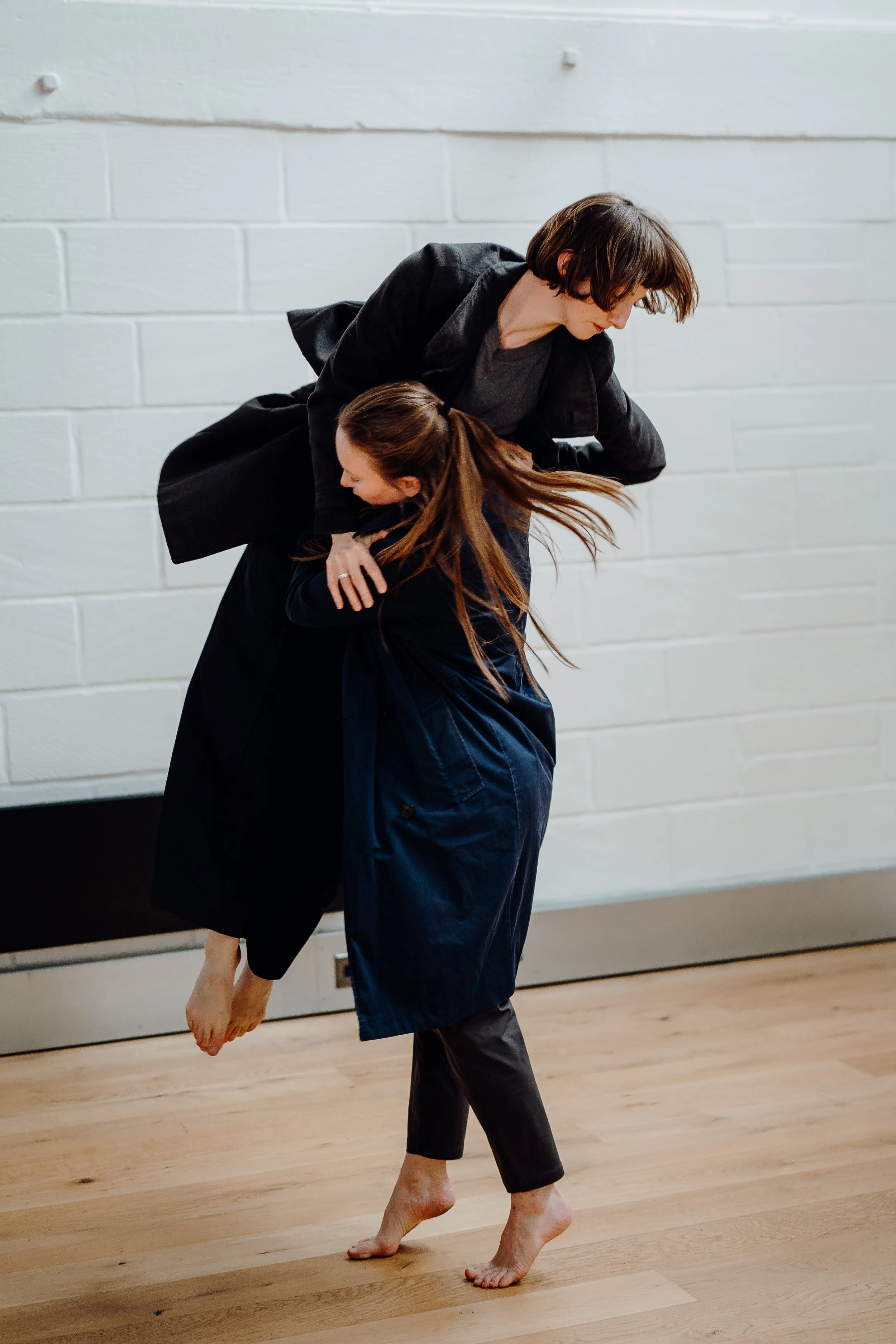 Two women practicing Aikido, with one woman lifting and turning the other in a martial arts move against a white brick wall.