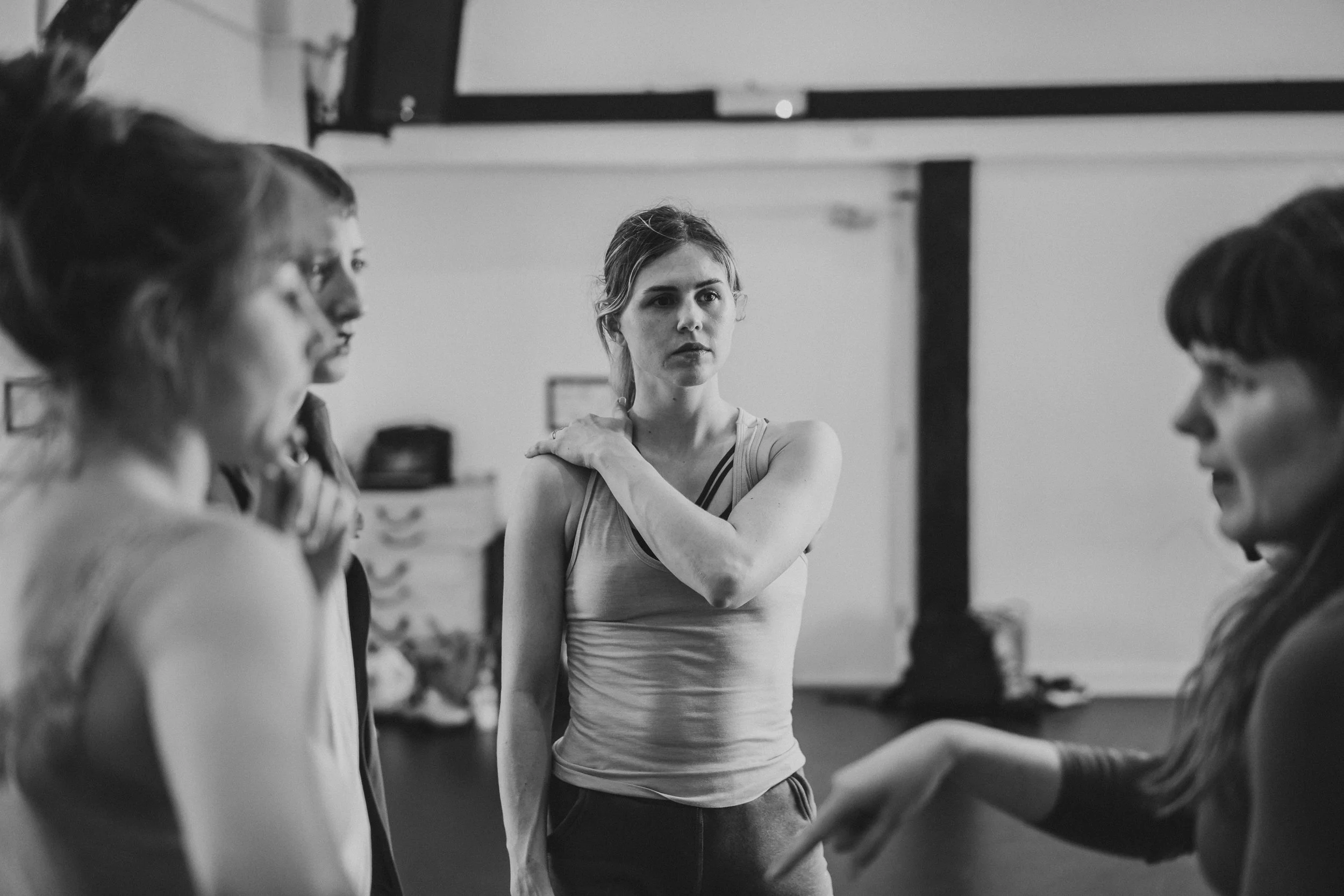 Black and white photo of a woman with a serious expression and her hand on her shoulder, surrounded by three other women in a dance studio or rehearsal space.