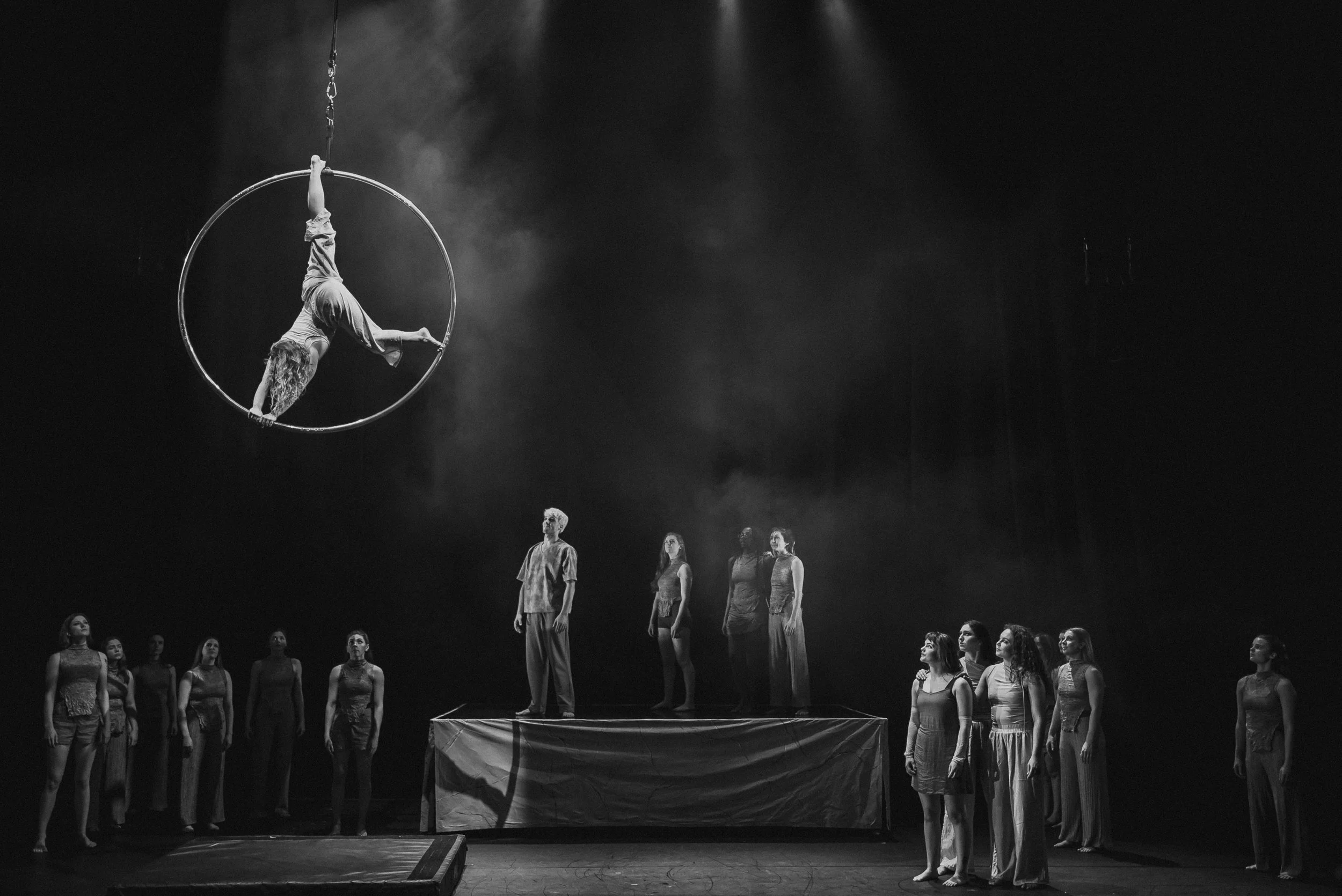 A black-and-white photo of a circus or acrobatic performance featuring a performer hanging upside down inside a large metal ring suspended from the ceiling, with a group of acrobats standing on a raised platform and on the floor beneath, watching.