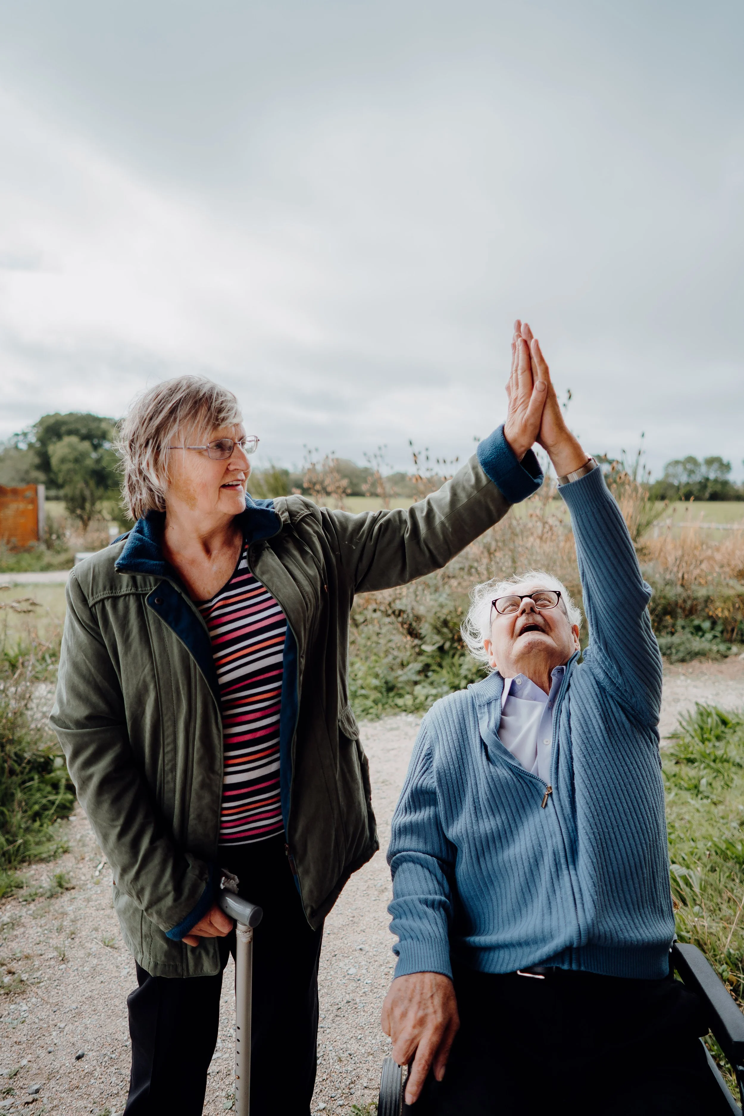 Two elderly women outdoors, one standing and holding a walking stick, the other sitting in a wheelchair, giving each other a high-five. They are smiling and surrounded by nature.