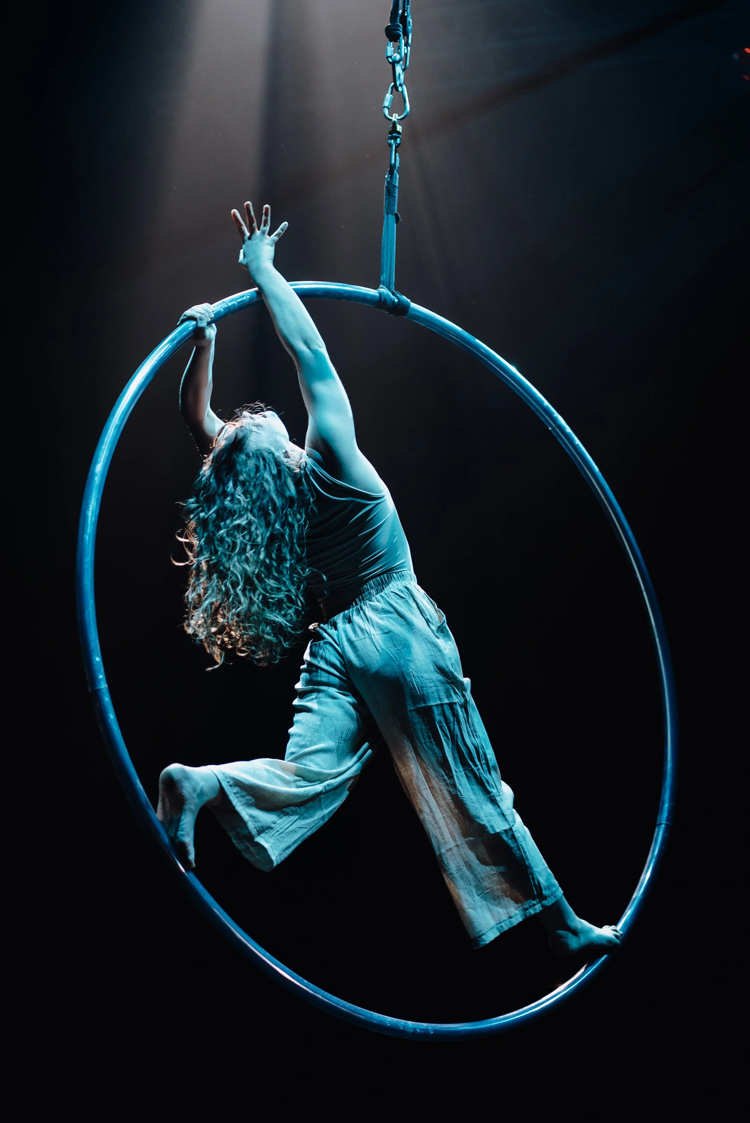 A woman performing an aerial acrobatics on a large hanging hoop, with one hand gripping the hoop and her body arched backward, set against a dark background with focused lighting.