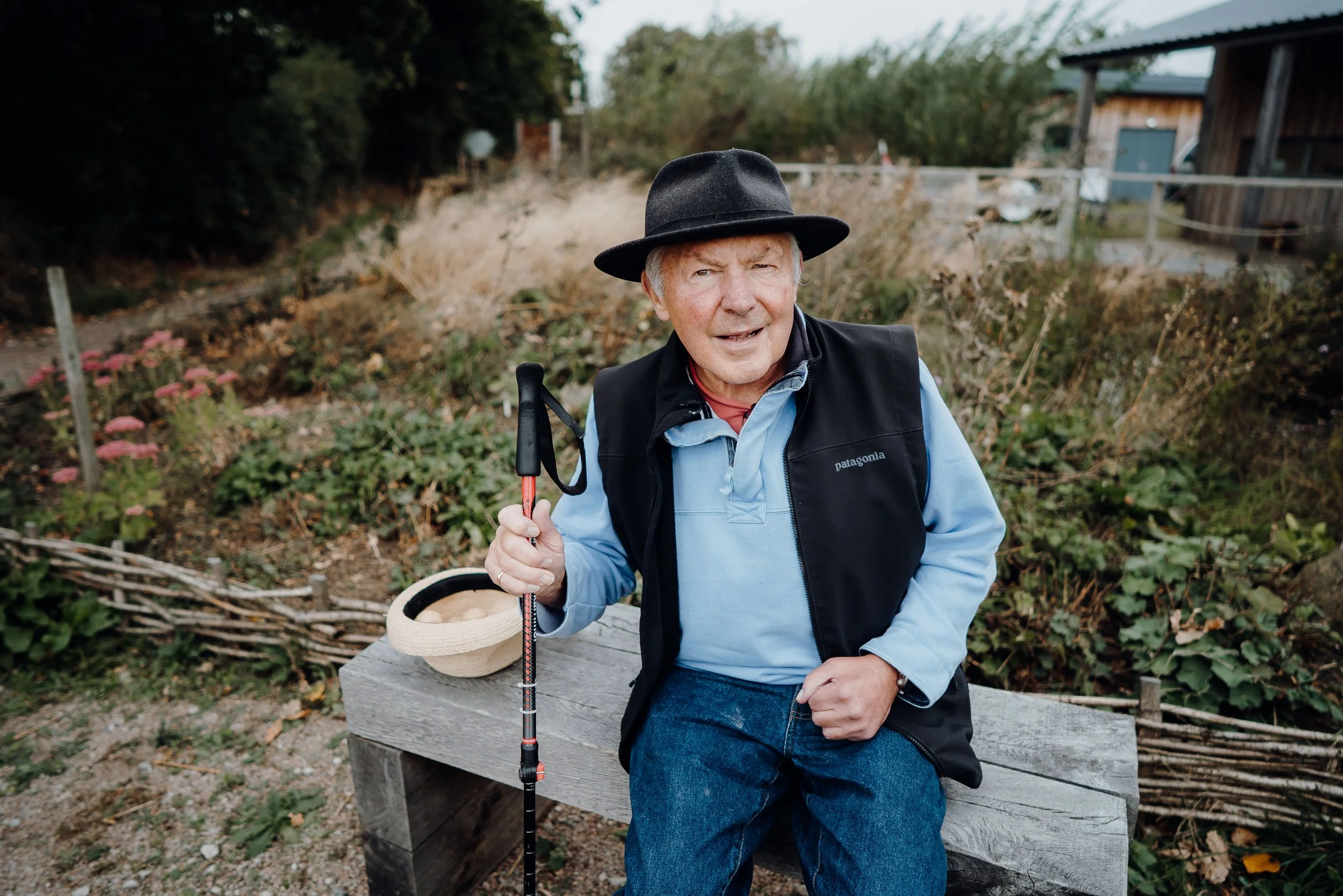 Elderly man sitting on a wooden bench in a garden, wearing a black hat and holding a walking stick.