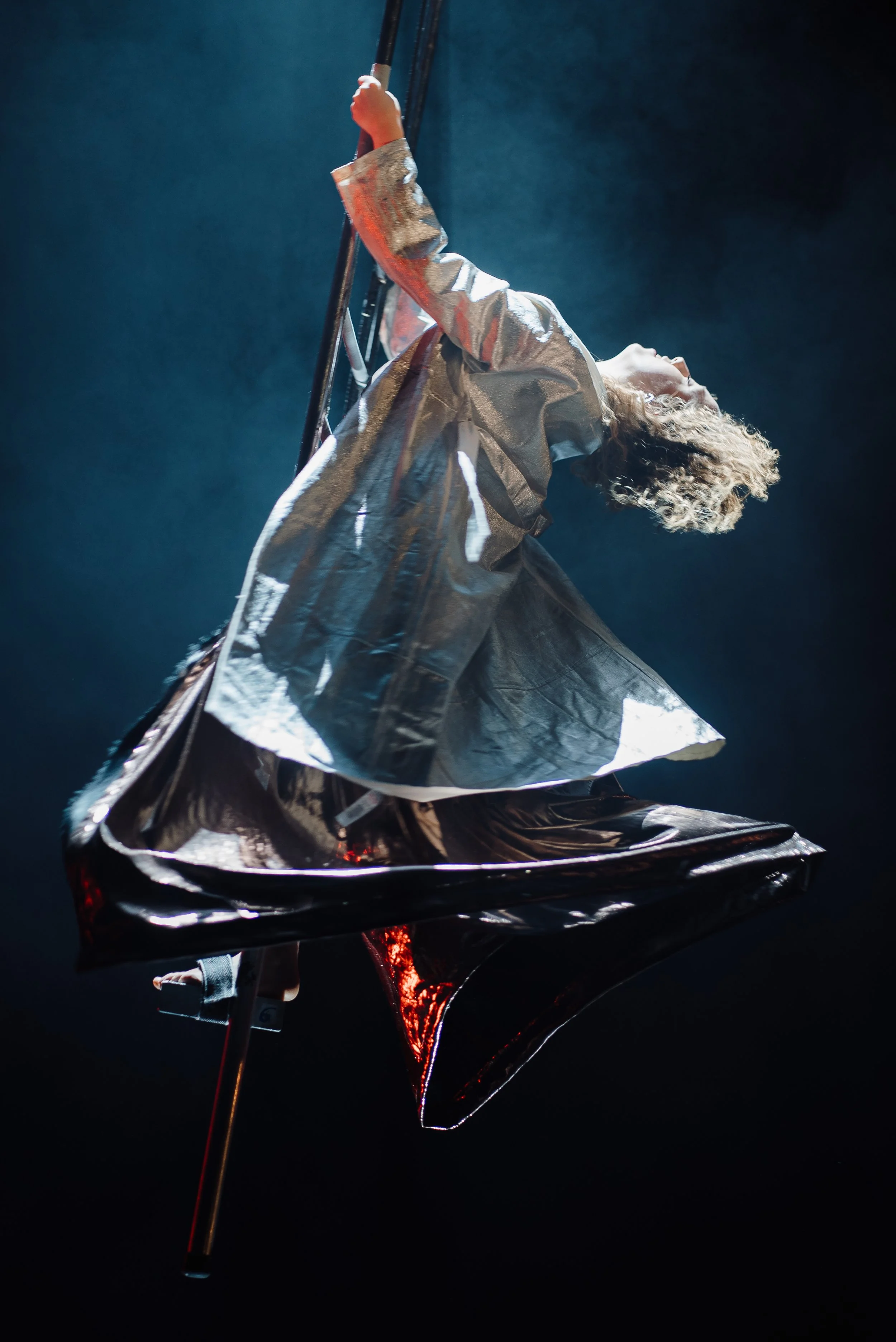 A performer on an aerial silk, dressed in metallic clothing, suspended in the air against a dark background with dramatic lighting.