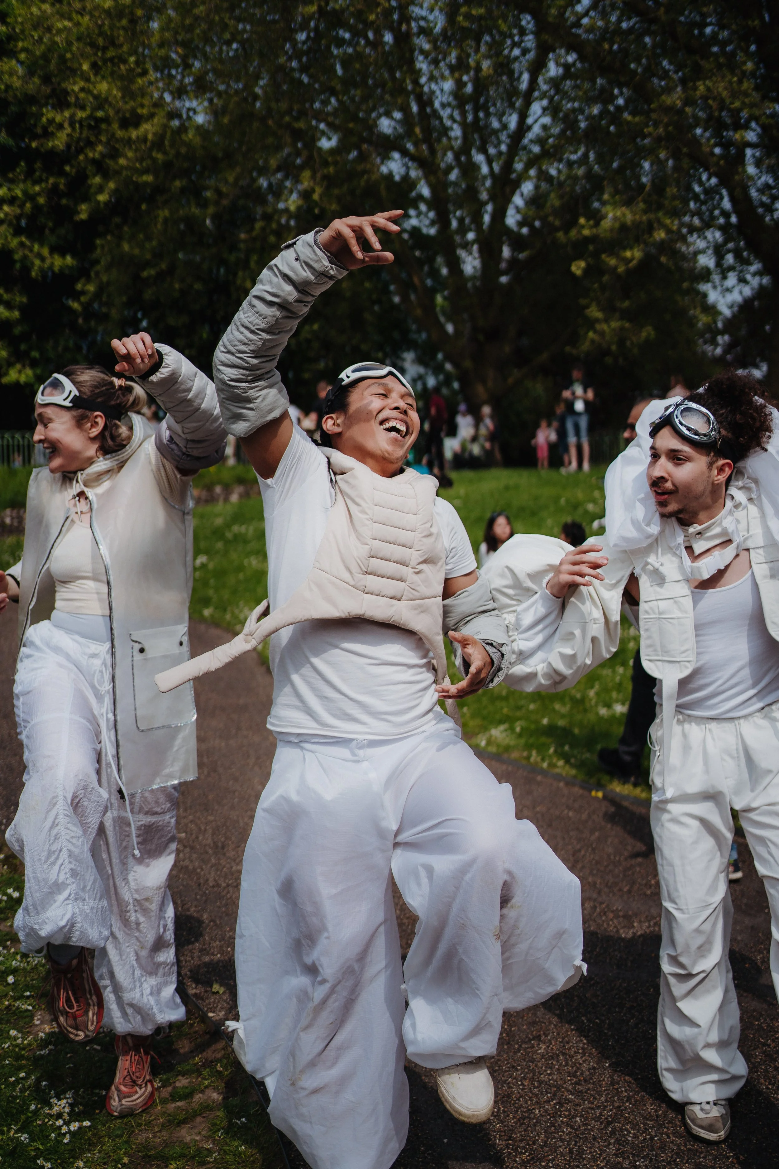 People joyfully dancing outdoors during daytime, wearing white and beige clothing, with some wearing goggles, on a park path with trees and other people in the background.