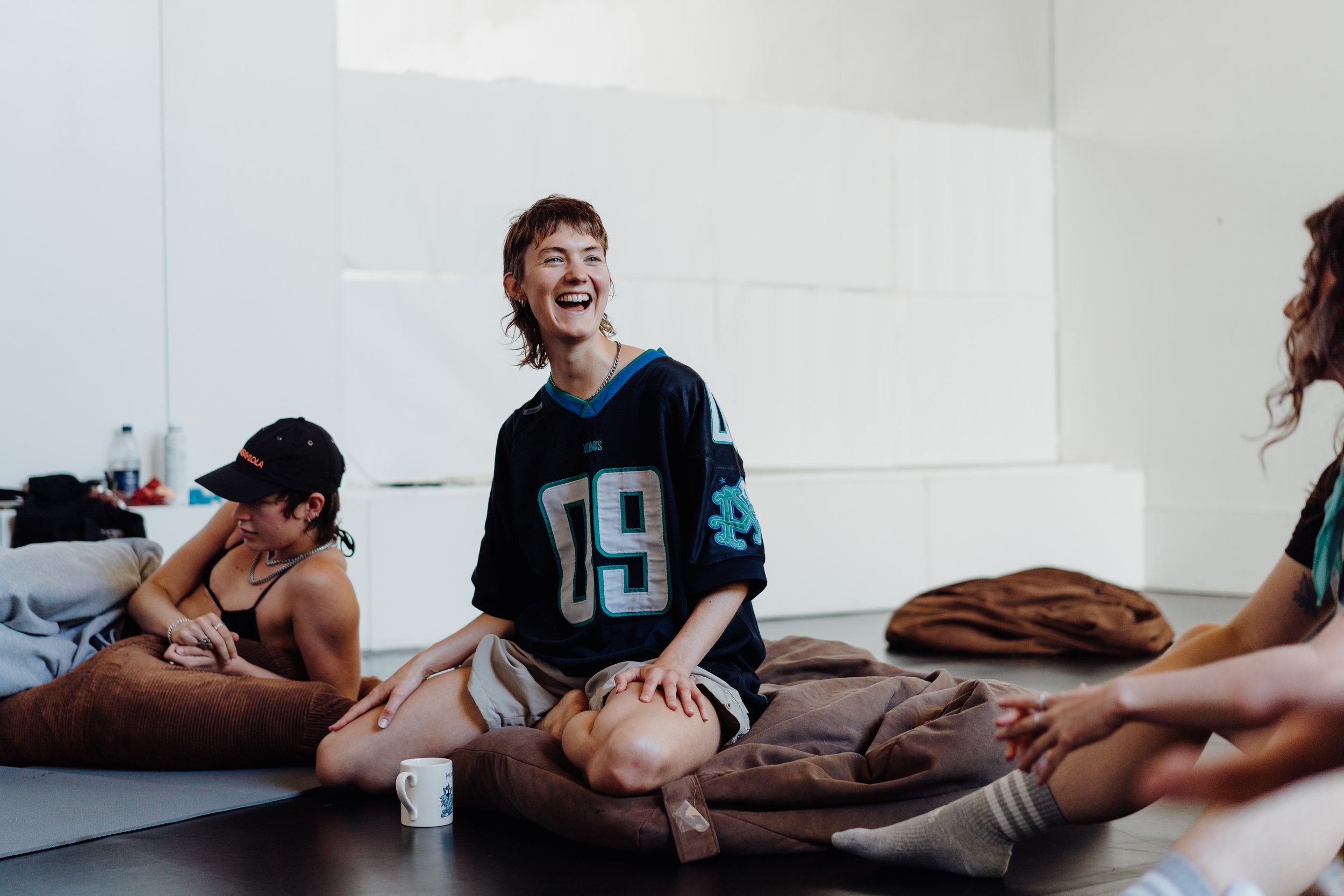 Group of young people sitting on mats and pillows, one in a black sports jersey with the number 19, smiling and laughing in a bright, white room.