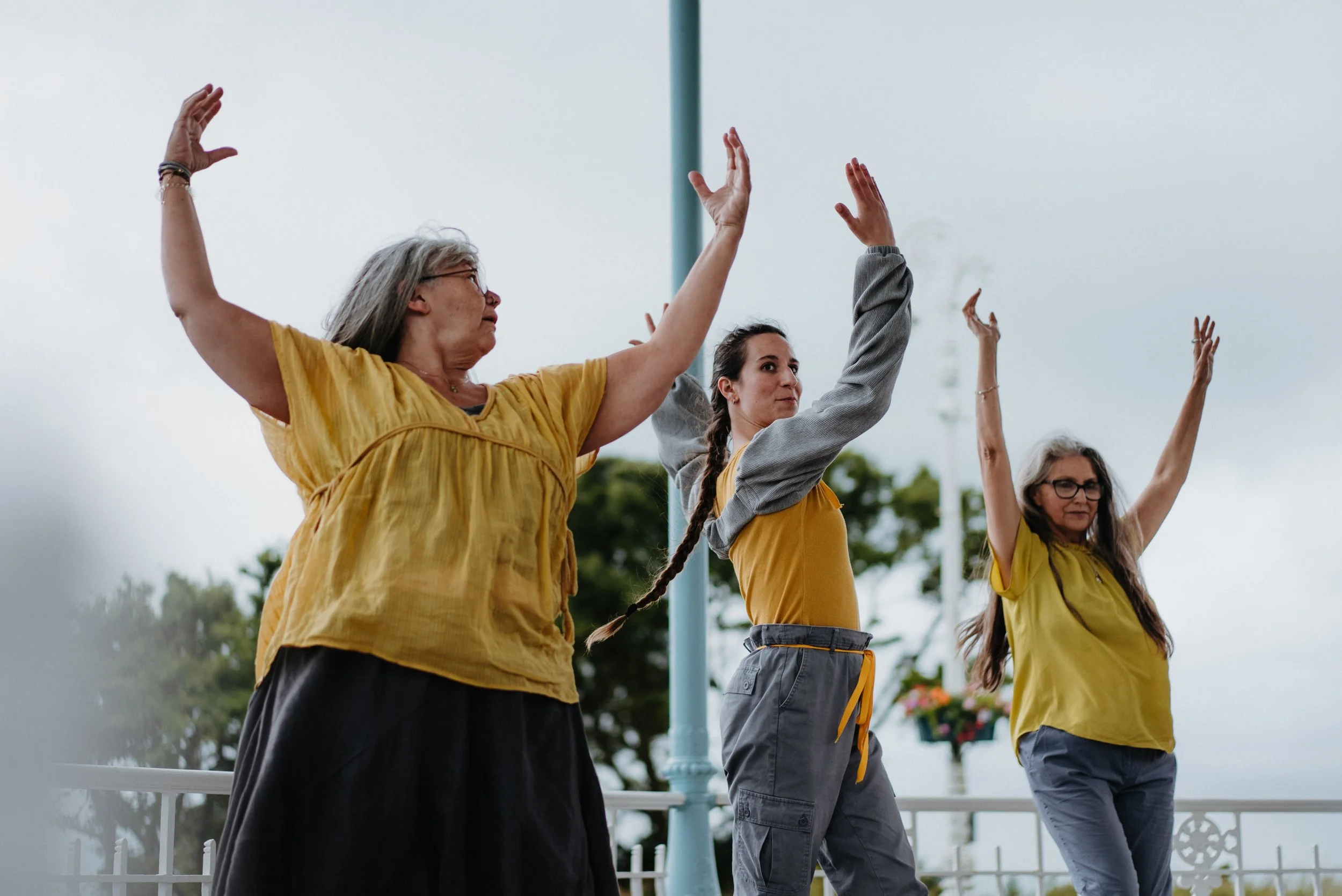 Three women standing outdoors, raising their arms in the air and smiling, with cloudy sky and trees in the background.