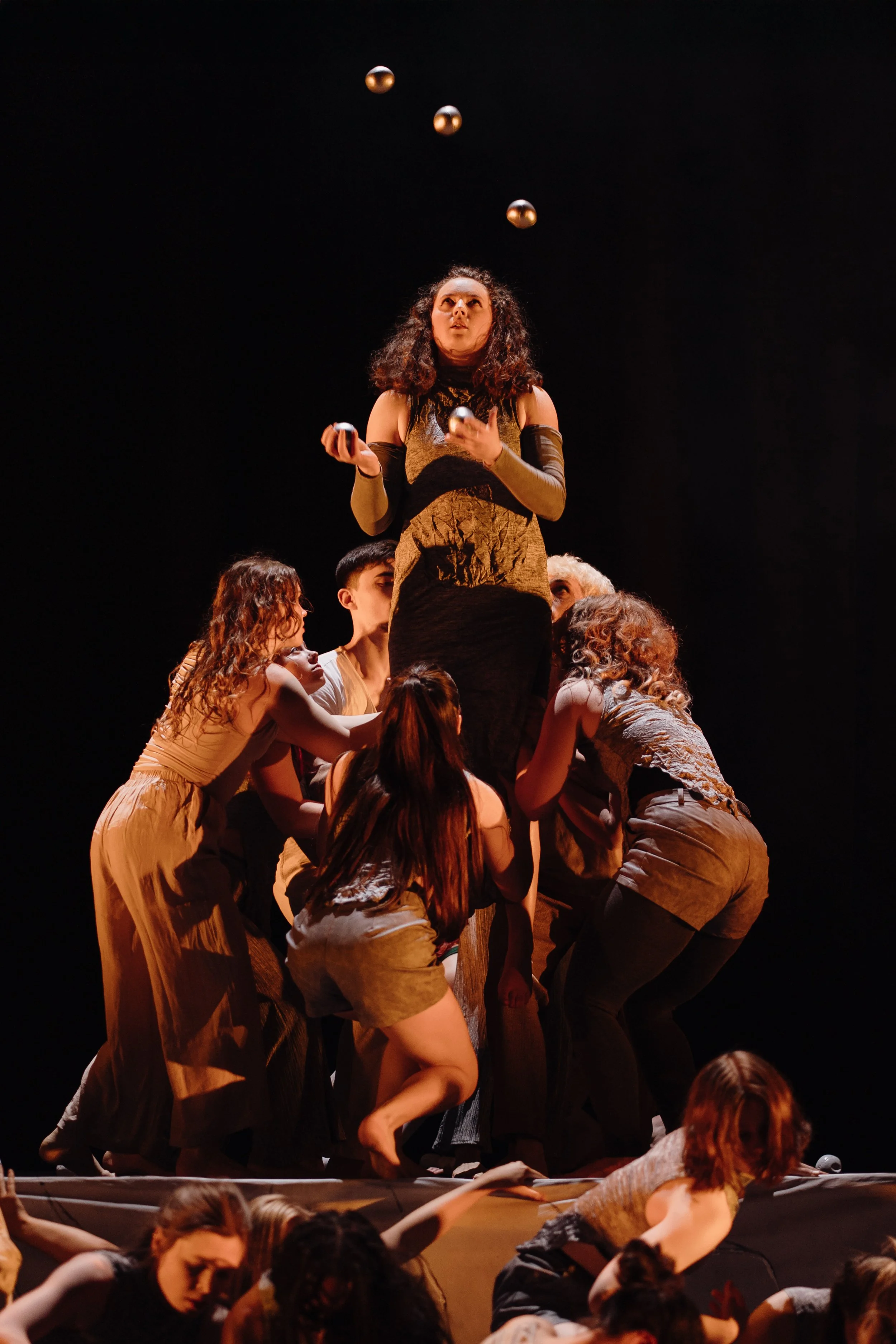 A woman standing on a platform with several people supporting her from below, juggling or tossing balls in the air during a stage performance.