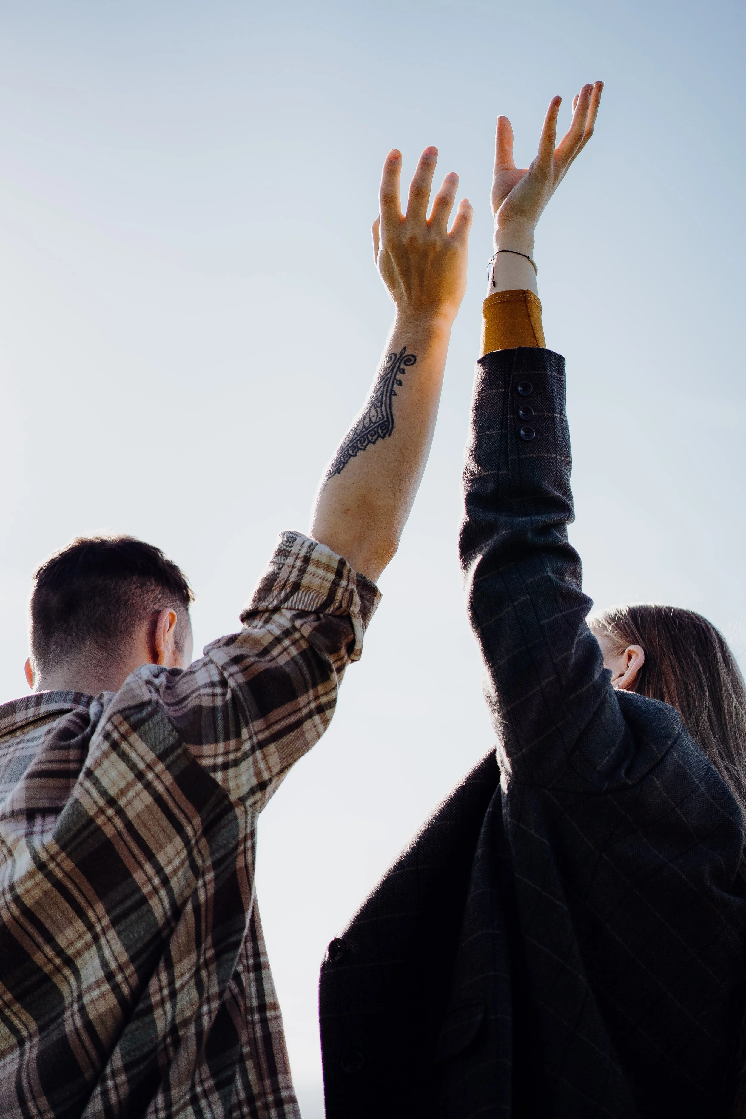 Two people raising their hands towards the sky outdoors during daytime, with one person wearing a plaid shirt and the other wearing a dark blazer.