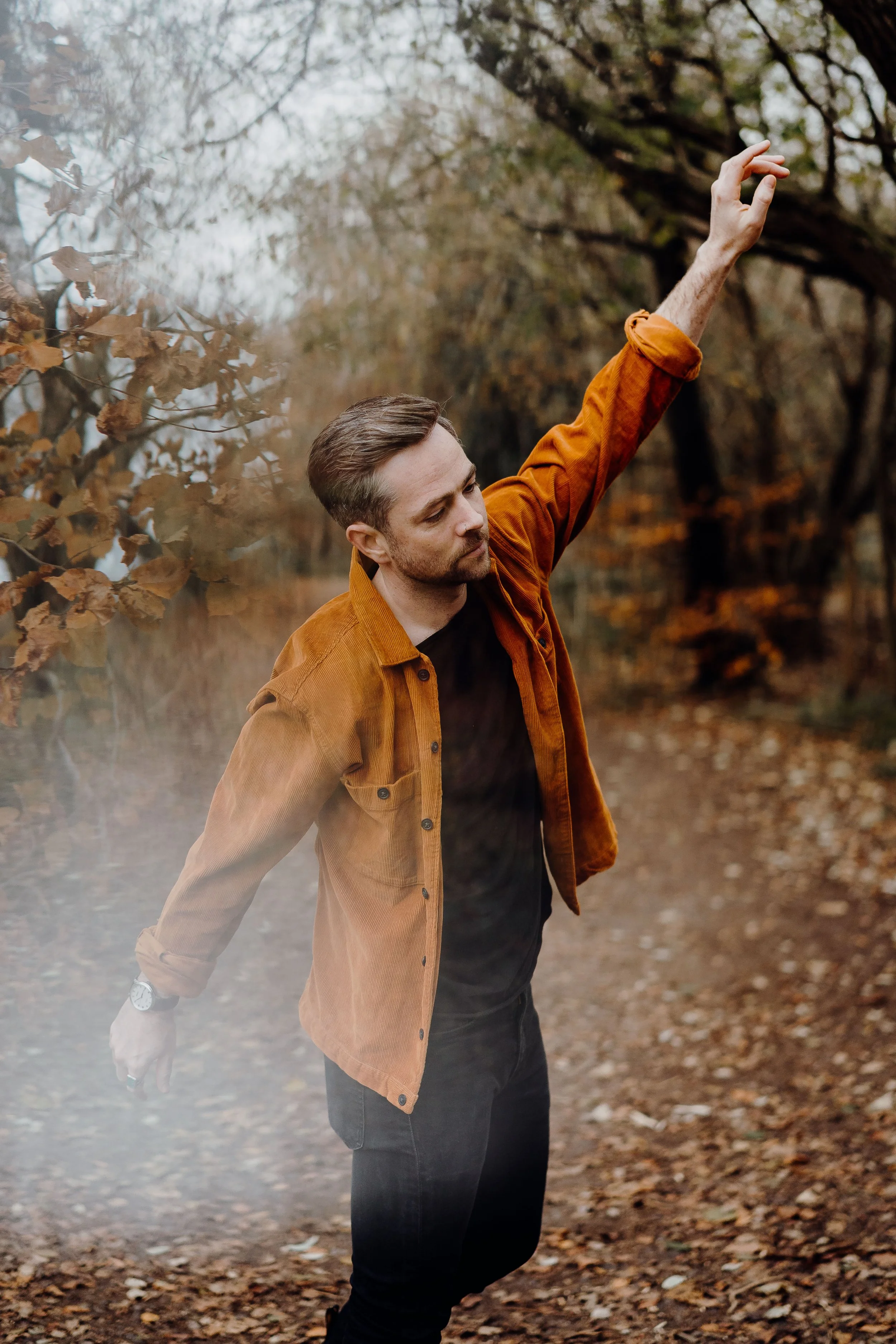 A man with brown hair and a beard, wearing an orange jacket over a black shirt, stands with closed eyes and one arm raised in a fall forest, with trees and leaves around him.