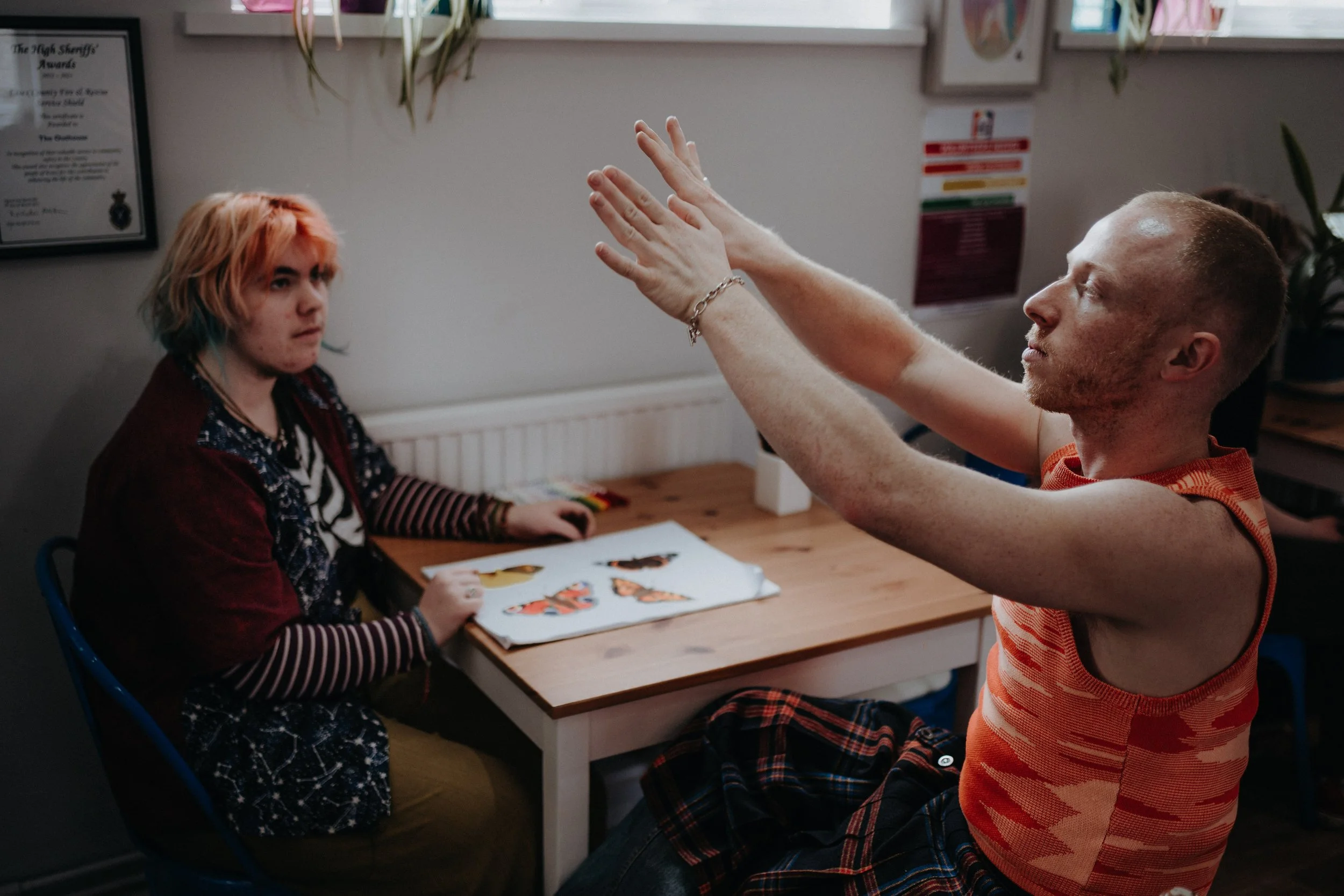 A man with tattoos on his arms, wearing an orange and red sleeveless shirt, raises his hands while talking to a woman with multicolored hair, sitting at a table with butterfly pictures, in a room decorated with plants and posters.