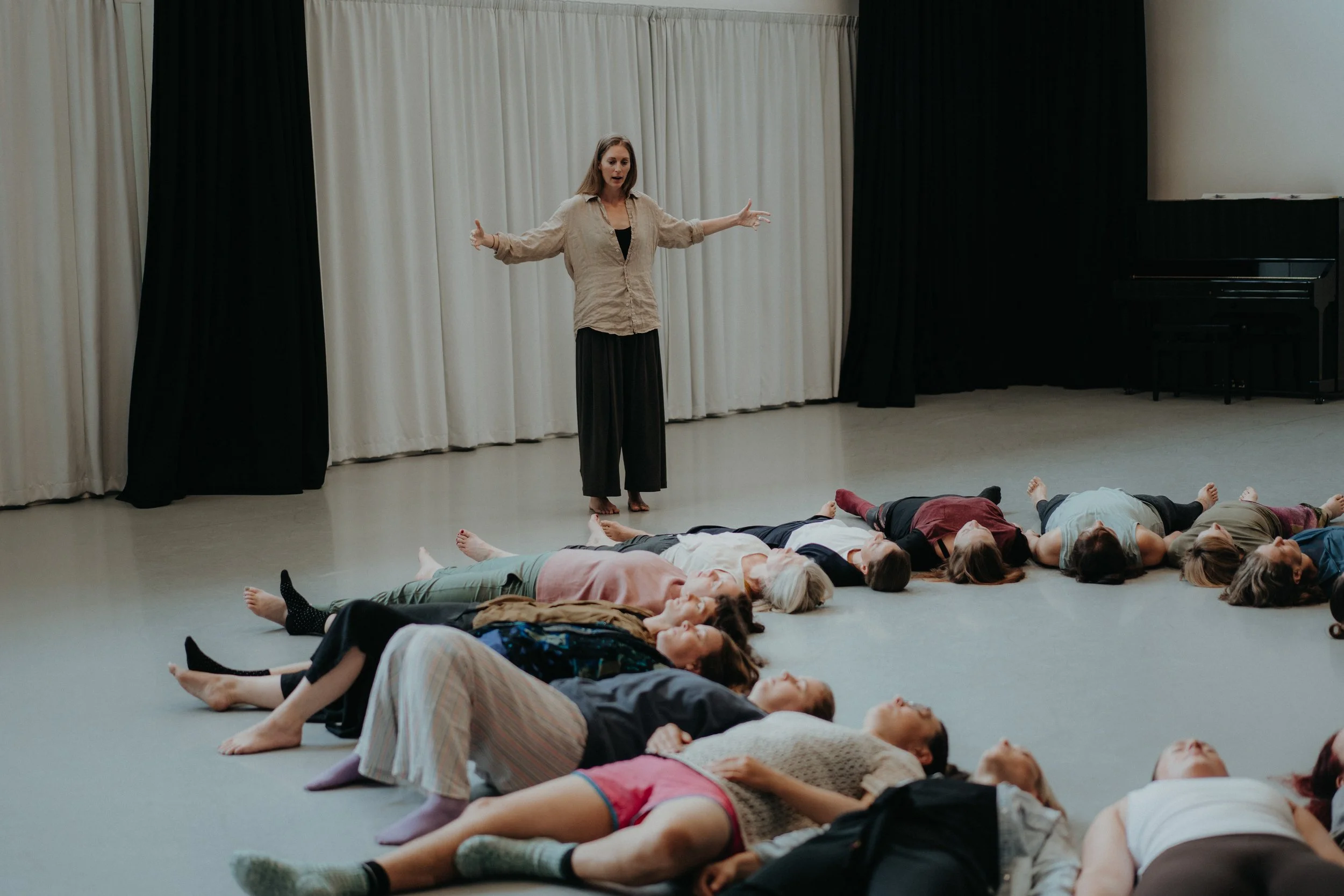A woman stands with her arms outstretched in front of a group of people lying on the floor, who are arranged in a semi-circle, in a room with black and white curtains and a piano in the background.