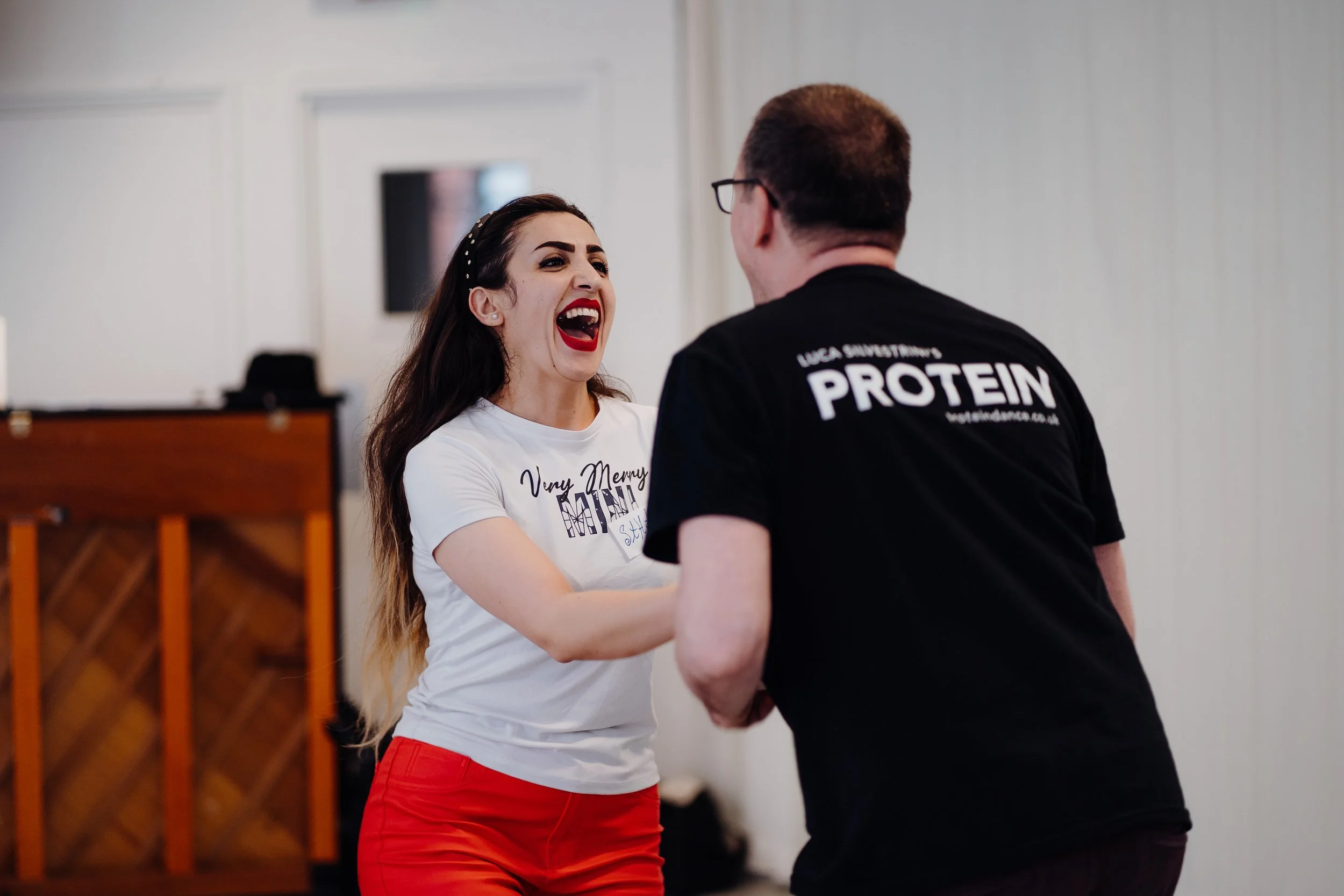 A woman laughing and dancing with a man in a black shirt that has "PROTEIN" written on the back.