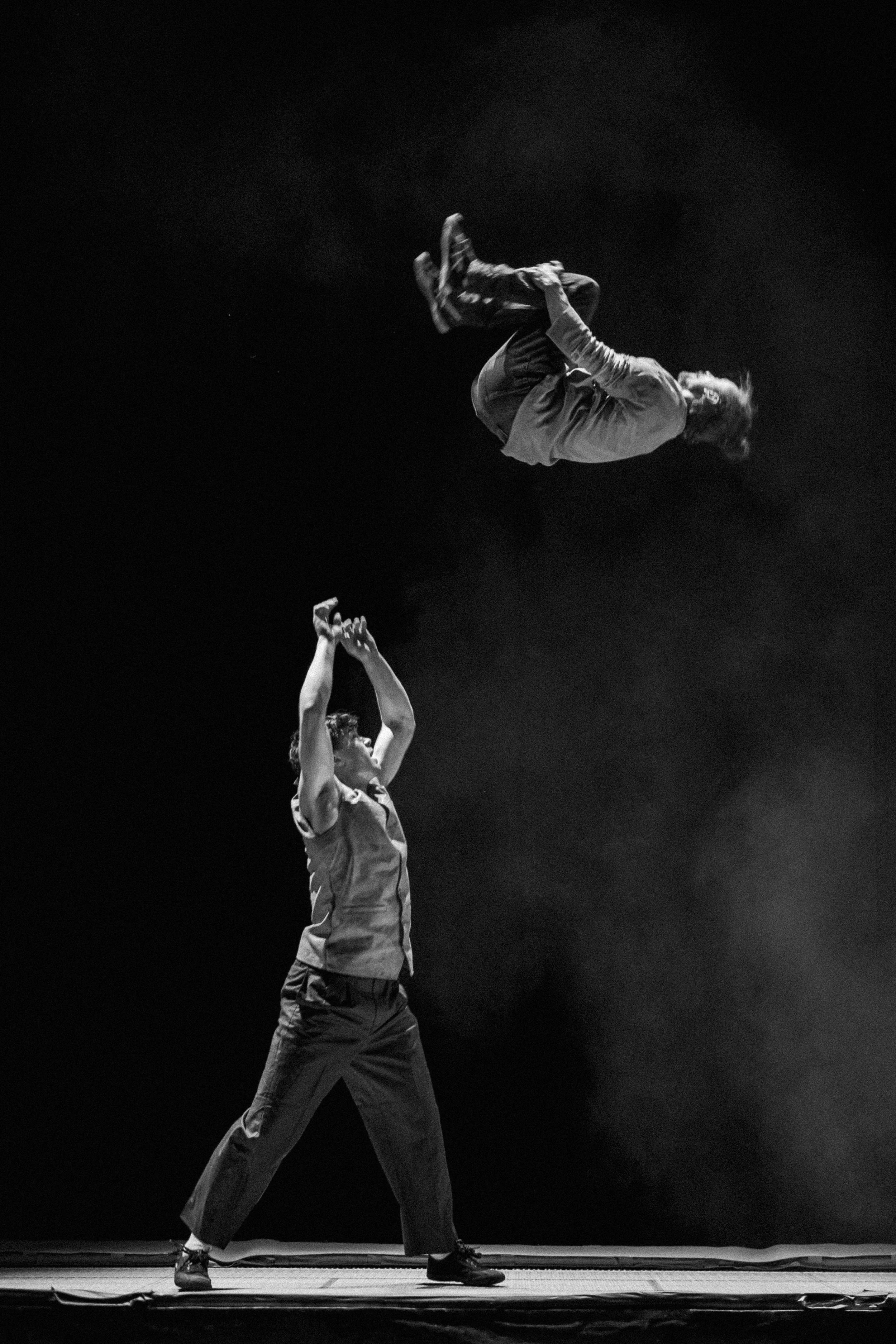 A black-and-white photo showing a performance where one person is standing on stage with hands raised, tossing another person into the air, against a dark background.