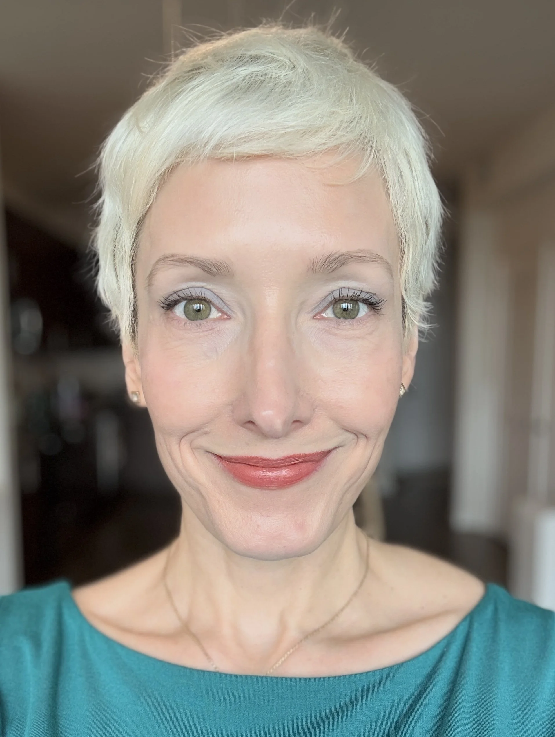 Close-up of a woman with short platinum blonde hair, light skin, green eyes, wearing subtle makeup, a teal top, and a delicate necklace, smiling slightly.