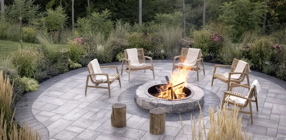 Modern Toronto backyard with stone patio, central fire pit, wood chairs, and native plants—creating a cozy outdoor space ideal for evening gatherings and natural landscape design