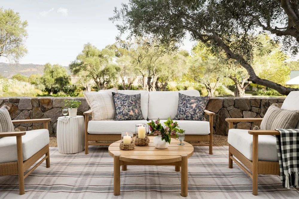 Outdoor patio with wooden furniture including a cushioned sofa, armchair, and round coffee table decorated with candles and flowers, set on a striped rug 