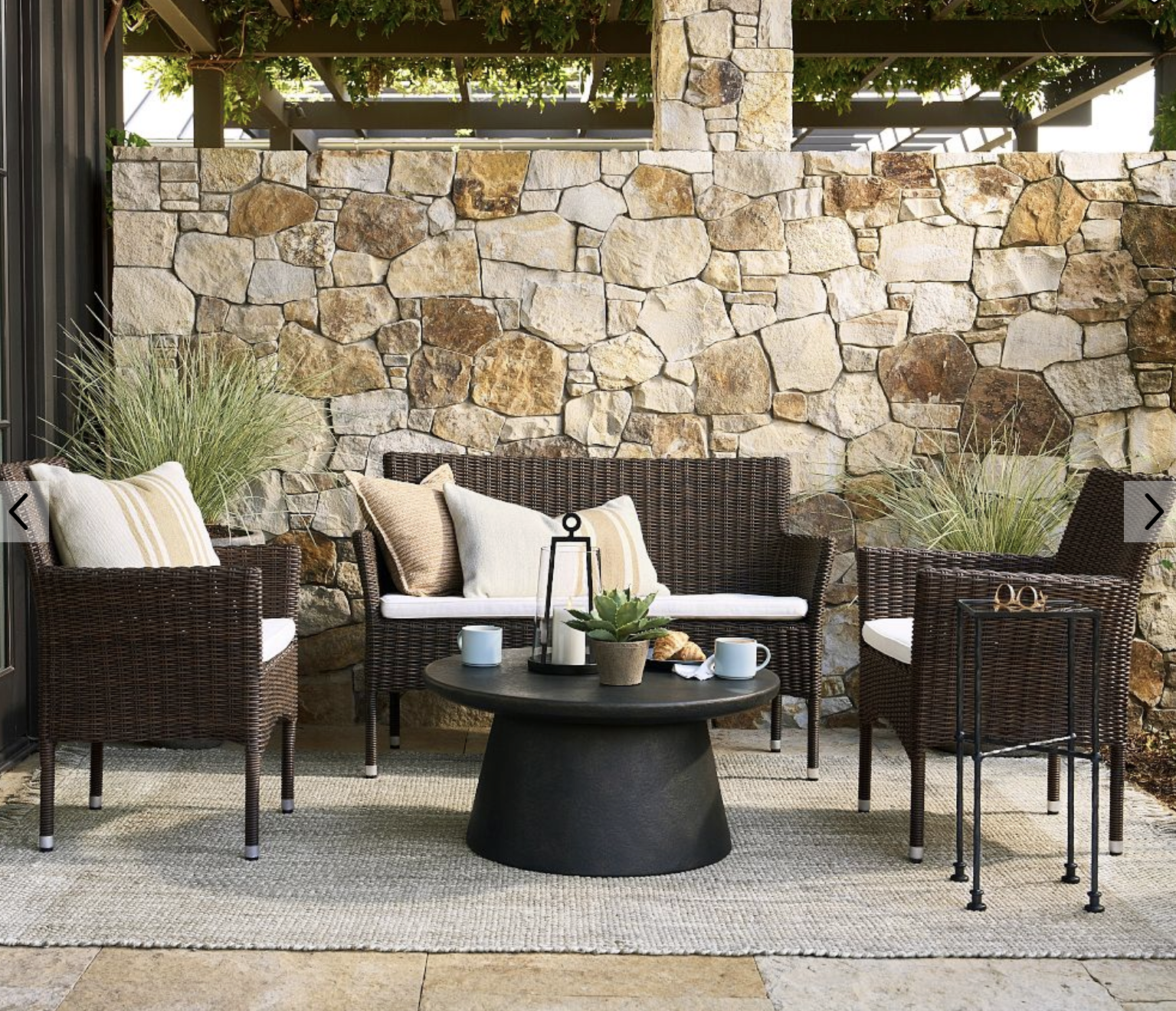 Outdoor patio with wicker chairs, a sofa, and coffee table, styled with neutral cushions and a stone wall backdrop.