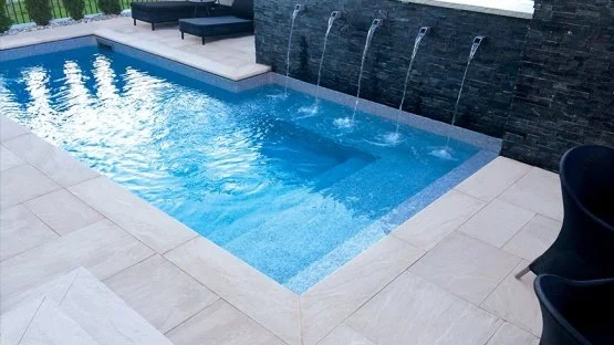 Swimming pool water feature with multiple wall spouts flowing into a pool framed by natural stone hardscape.