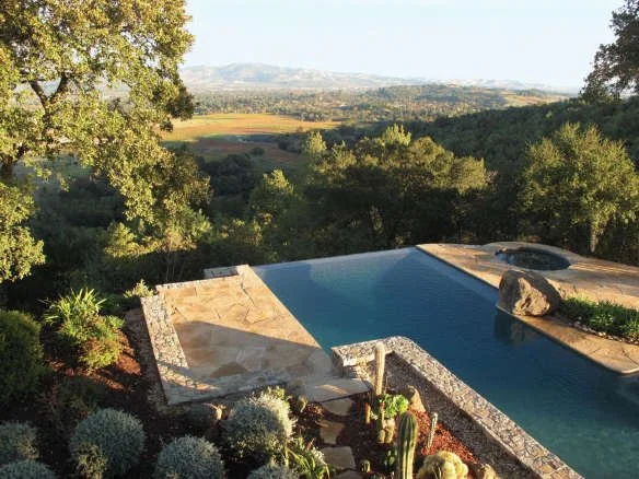 Infinity-edge swimming pool with natural stone terrace overlooking a scenic landscape and surrounding vegetation.