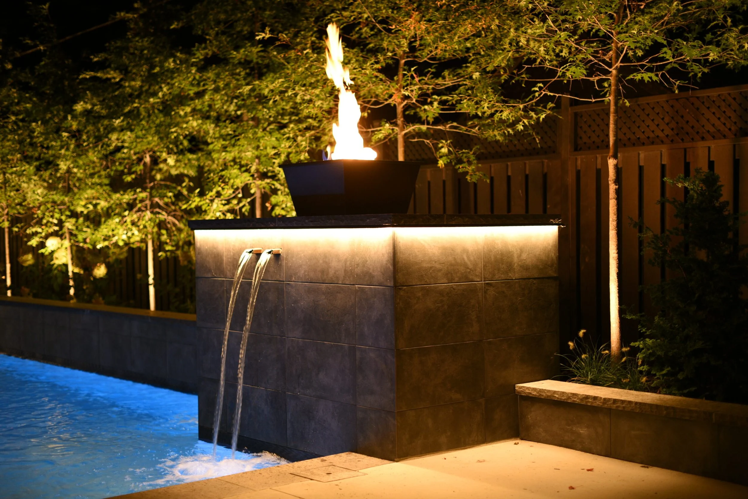 Modern fire and water feature integrated into a natural stone wall, creating a dramatic focal point in a luxury Toronto backyard landscape design at night.