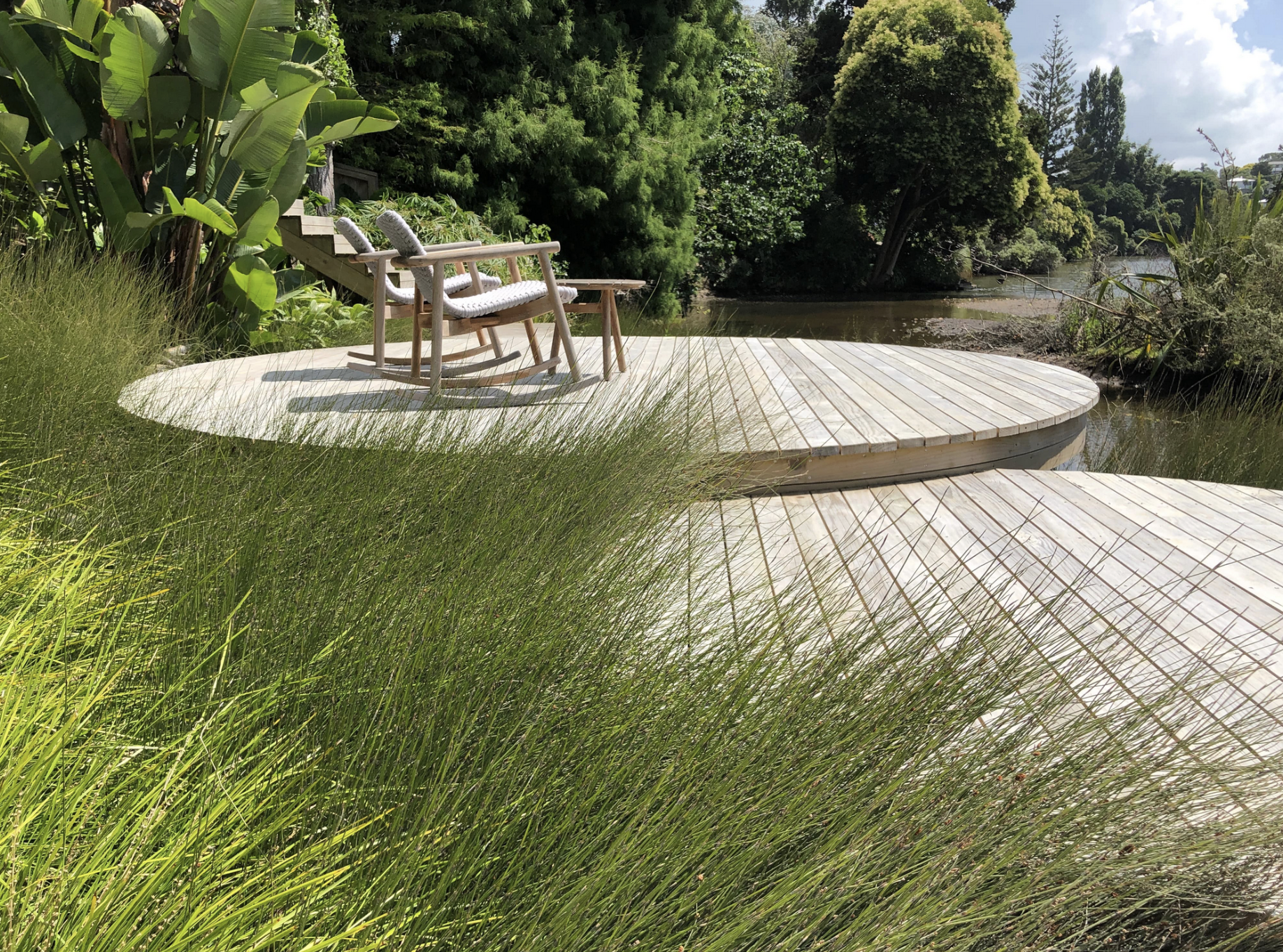 Circular wood deck platform surrounded by ornamental grasses with two lounge chairs in a natural garden setting.