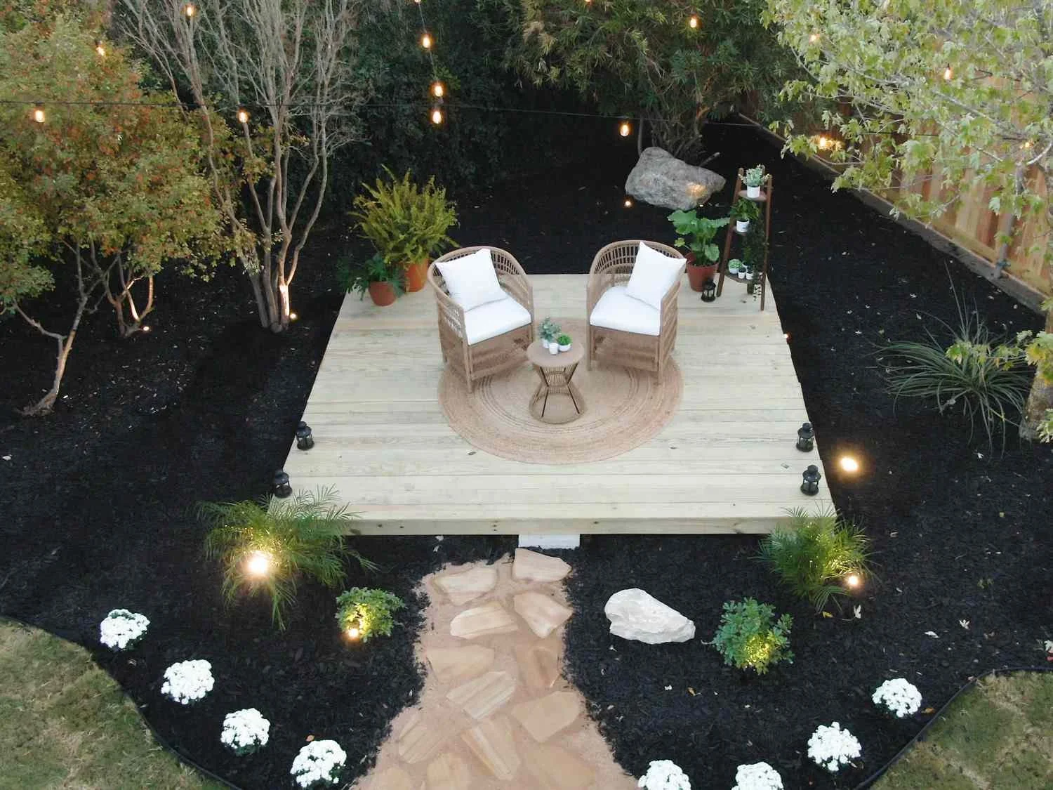 Small raised backyard deck with two chairs, soft landscape lighting, dark mulch garden beds, and stepping stone pathway.