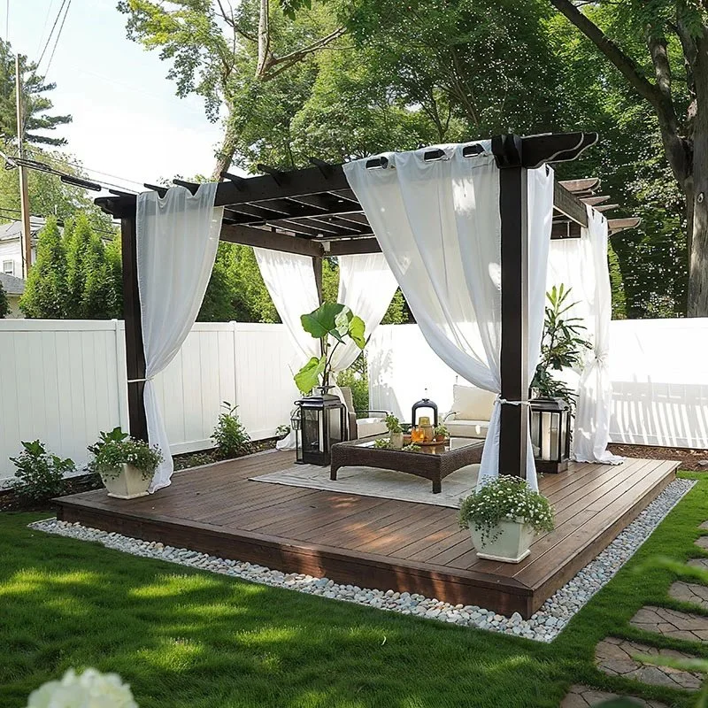 Pergola-covered backyard deck with white drapes, outdoor seating, and lush green lawn for a cozy outdoor living space.