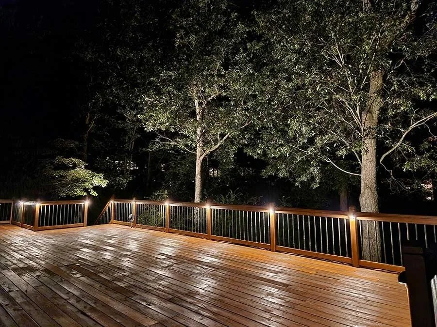 Spacious wood deck at night with railing post lights highlighting surrounding mature trees.