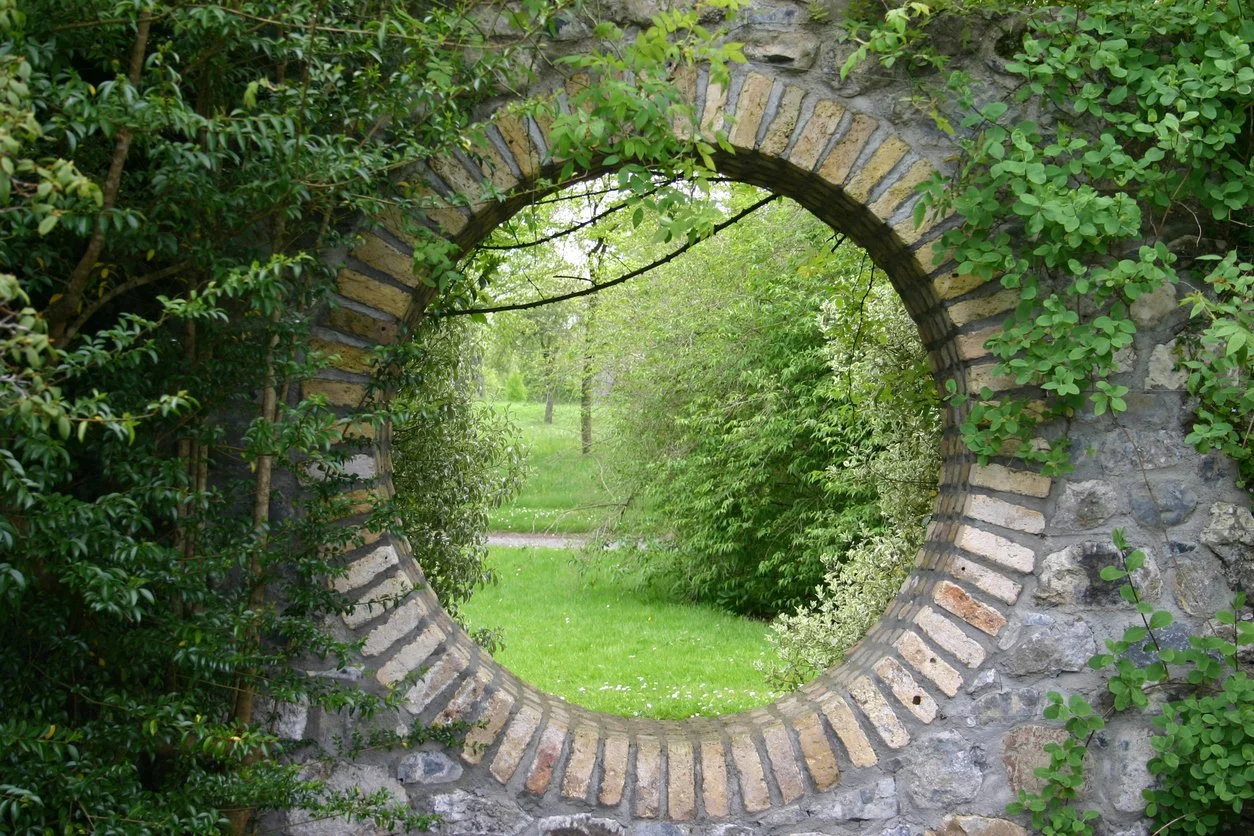 A circular brick and stone wall opening surrounded by green leafy plants and trees, revealing a lush grassy garden area.