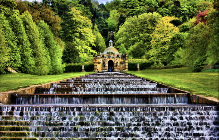 Water cascading over tiered terraces leading to a historic building with a dome, surrounded by lush green trees.