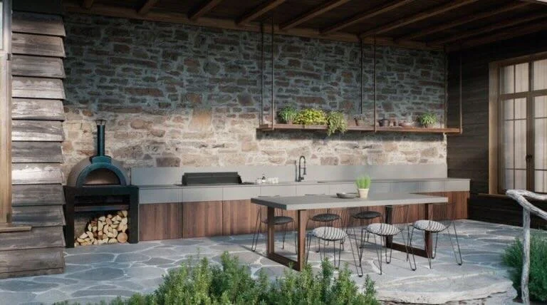 Outdoor kitchen with a brick wall, wood accents, and a seating area with four white chairs and a wooden table. There is a wood-fired pizza oven and a counter with a sink. Green plants are on a shelf above the counter, and there's a window with sunlight coming through.