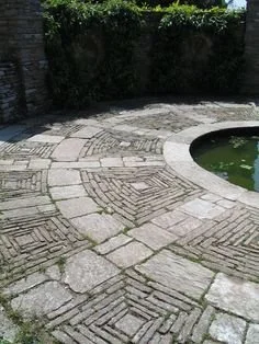 A stone paved patio area with a small round pond, surrounded by lush green bushes and stone walls.