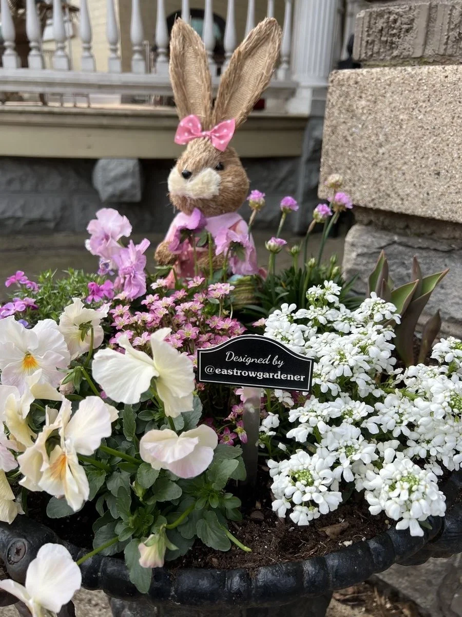 A decorative bunny figurine with long ears wearing a pink bow and dress, placed among pink and white flowers in a black planter, with a sign that says 'Designed by @eastowgardener'.