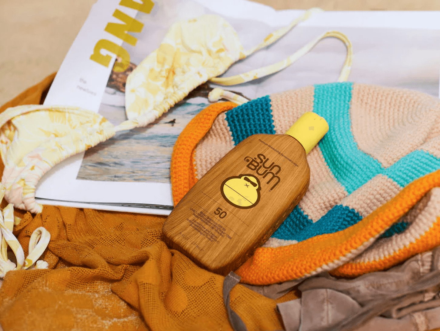 Sun Bum's Original SPF 50 Sunscreen Lotion laying on beach towel with bag, bikini, and magazine