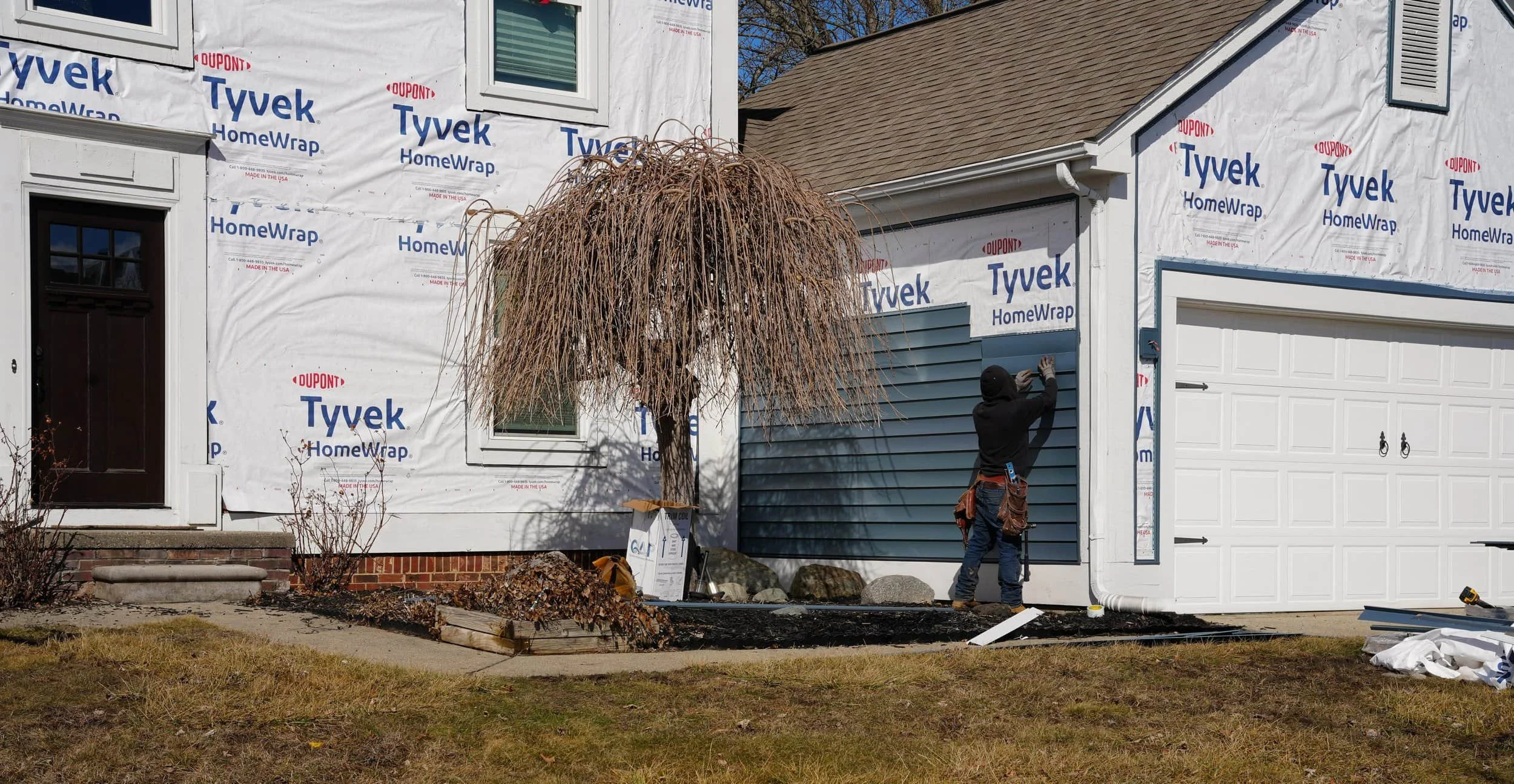A worker installs blue James Hardie siding on a house exterior with Tyvek wrap, next to a garage and a leafless tree in the yard.