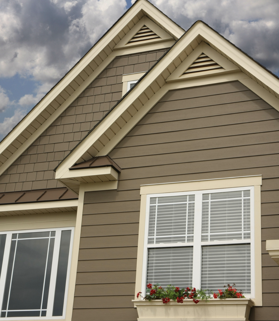 Exterior of a tan house with white trim, featuring a window with blinds and a flower box with red flowers. The house has pitched roofs, and the sky is cloudy.