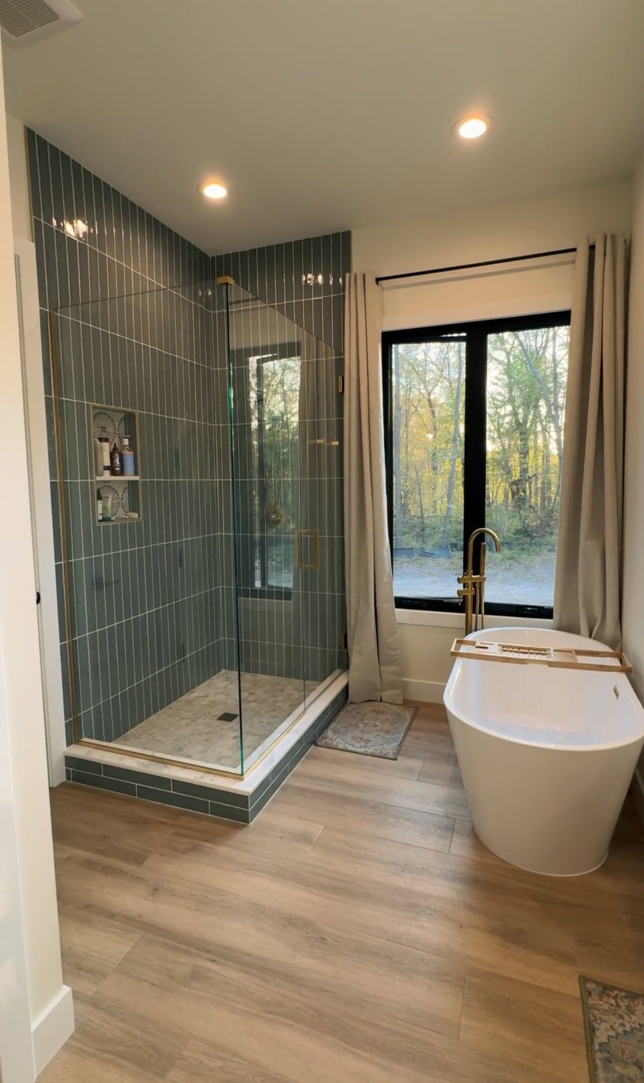 A modern bathroom remodeling featuring a glass-enclosed shower with dark green tiles, a large window with white curtains, and a freestanding bathtub with a gold faucet. The room has wood-look flooring and is well-lit with recessed ceiling lights.