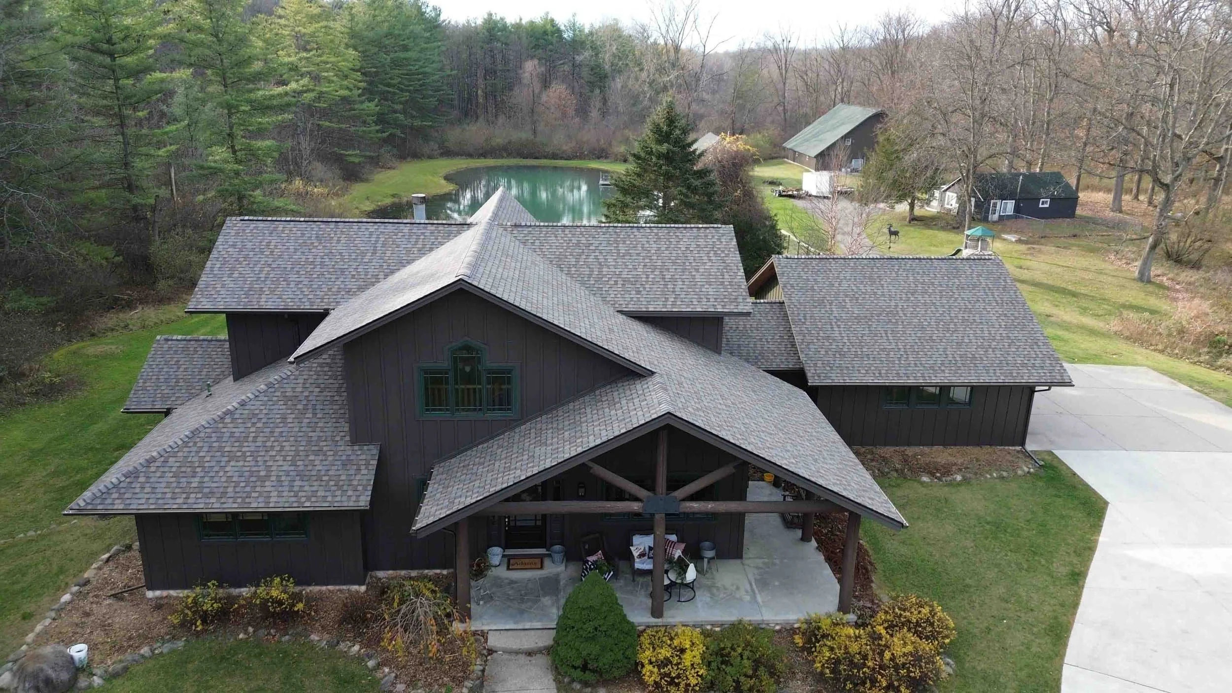 Aerial view of a brown house with a complex roof design, surrounded by greenery and trees, next to a small pond in a rural setting.