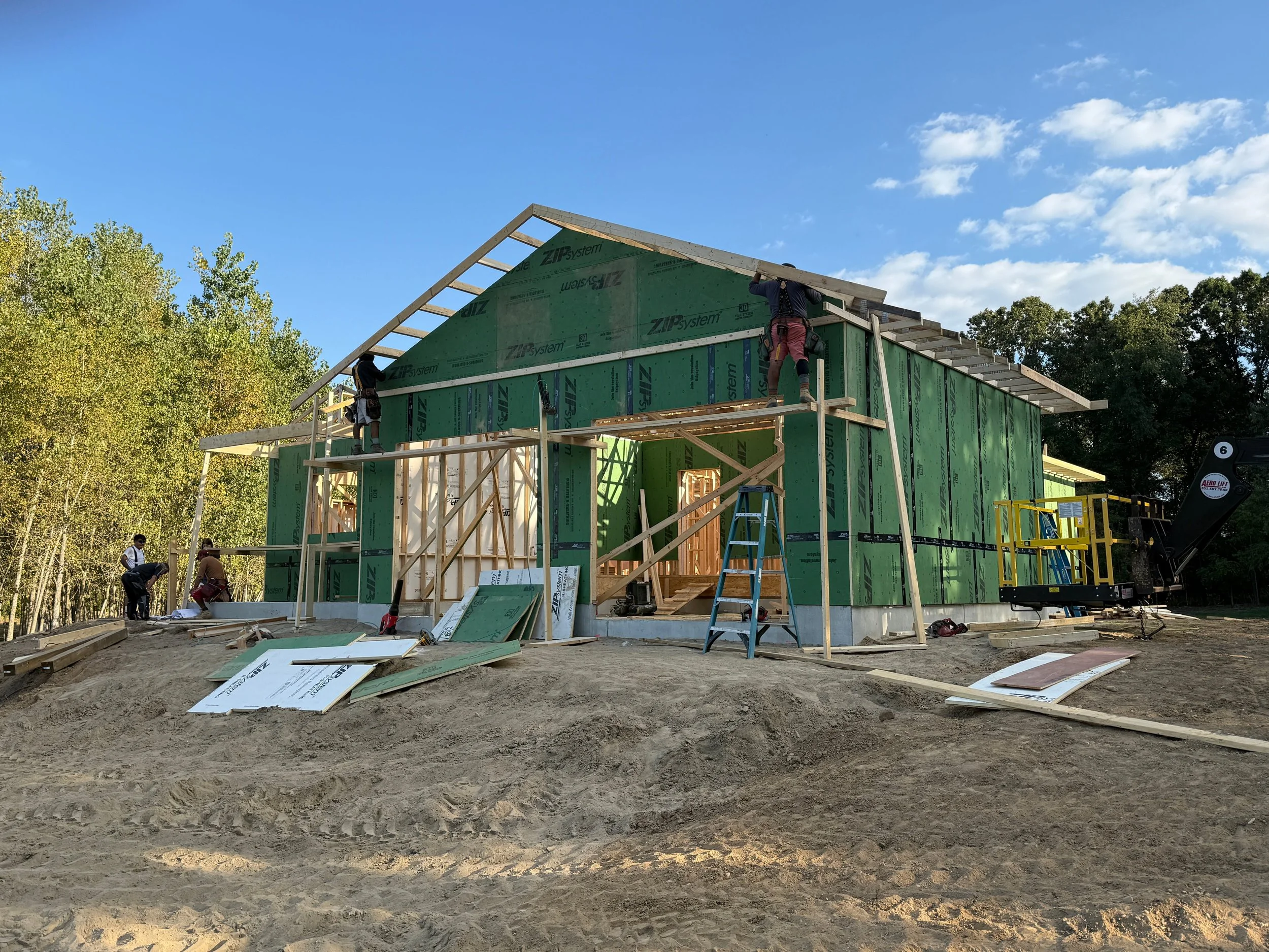 Construction workers building a house with green sheathing during daylight, with trees and a partly cloudy sky in the background.