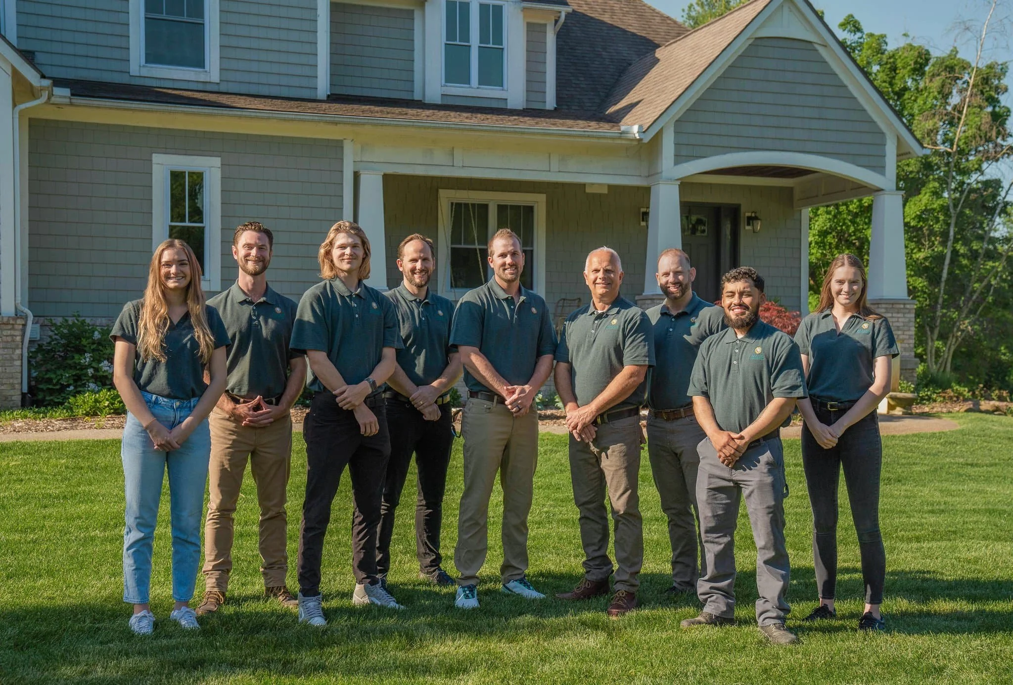 Eight people stand in a row on grass in front of a house, all wearing matching dark green shirts and smiling at the camera.
