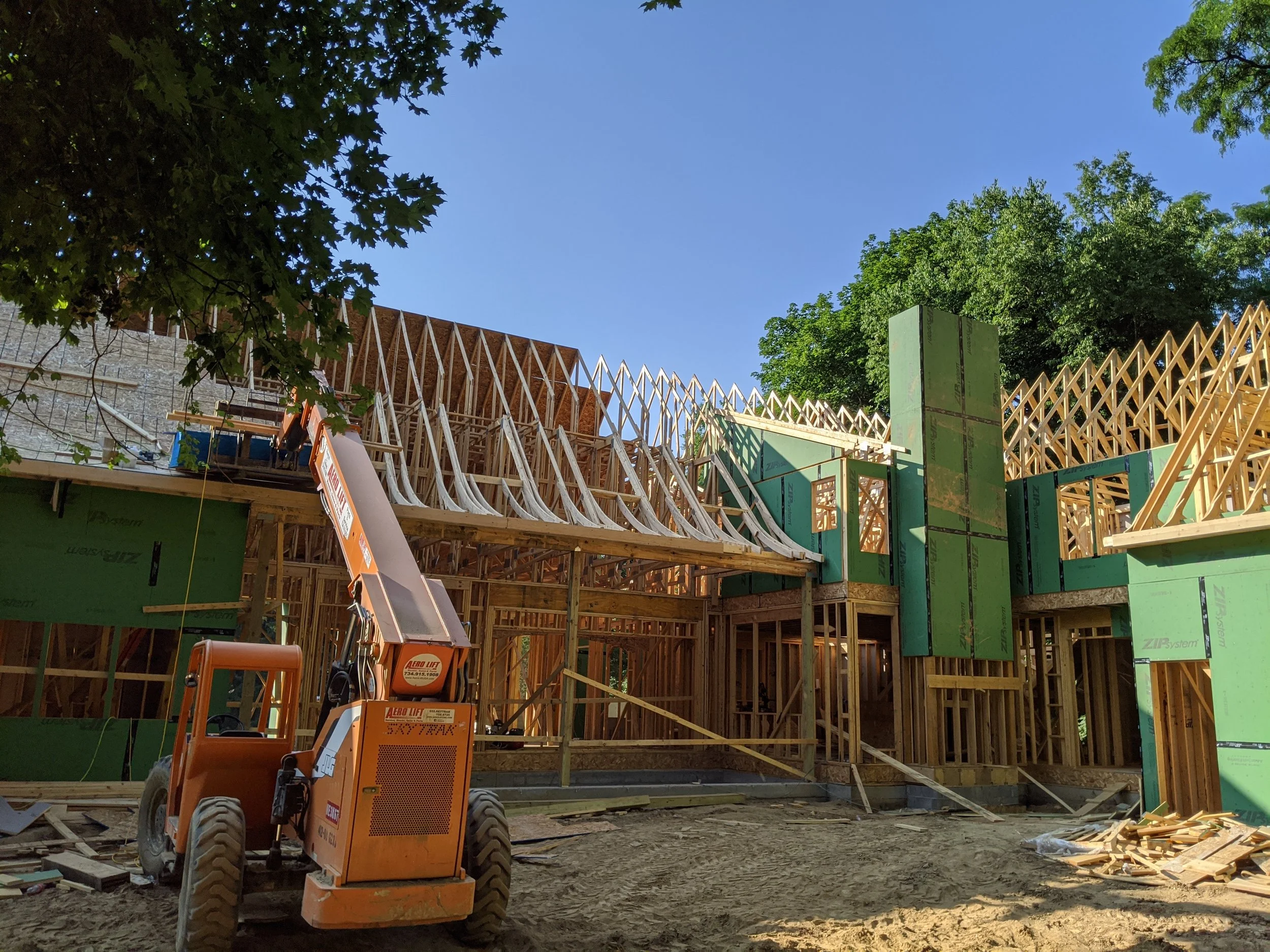 Construction site of a wooden house frame with partially built walls and roof, construction equipment, and green insulation panels, under a blue sky with green trees in the background.