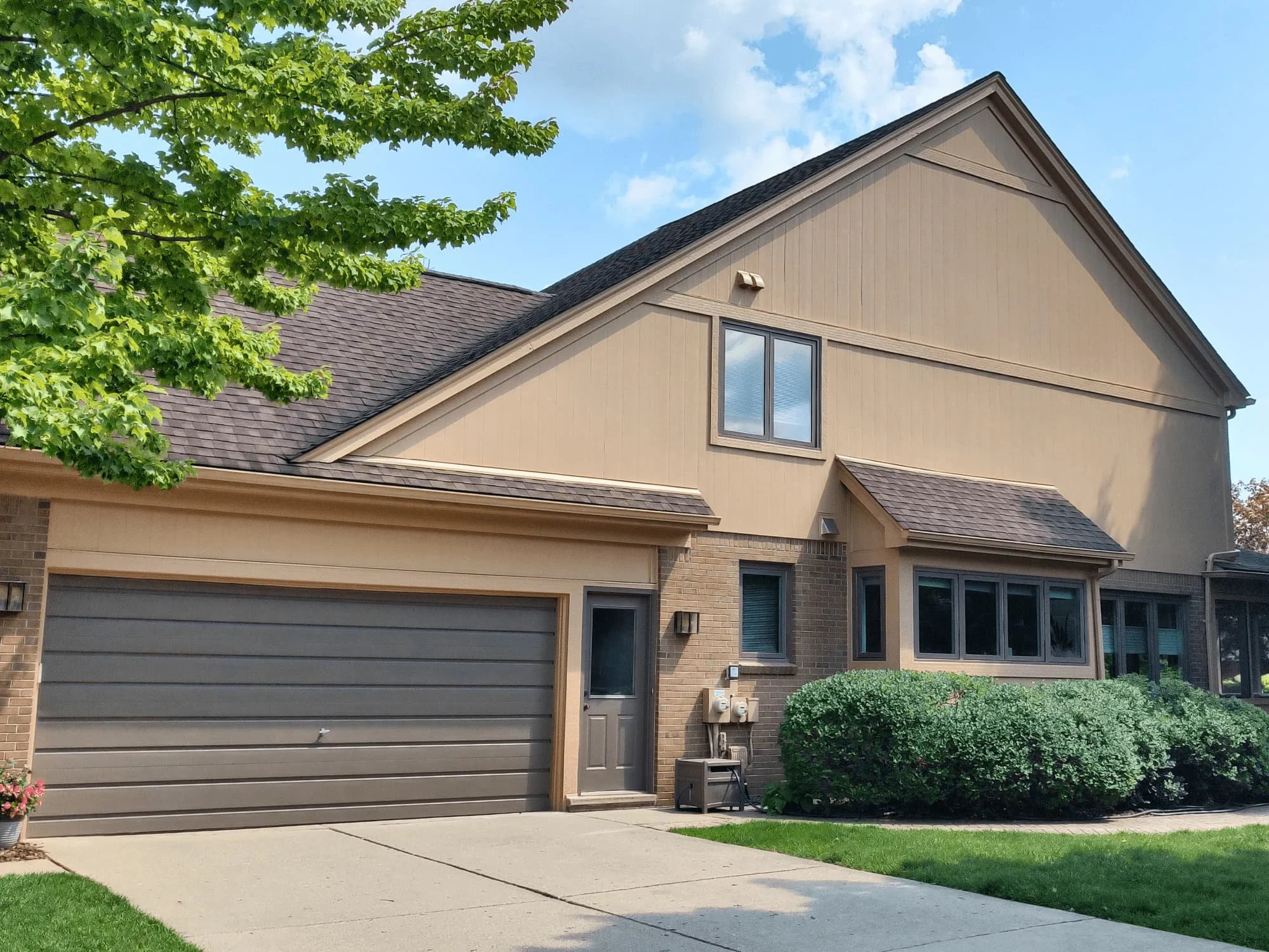 Two-story suburban house with beige siding and brick accents, featuring a large garage door. Surrounded by green lawn and shrubbery, with a tree partially visible on the left. Clear blue sky in the background.
