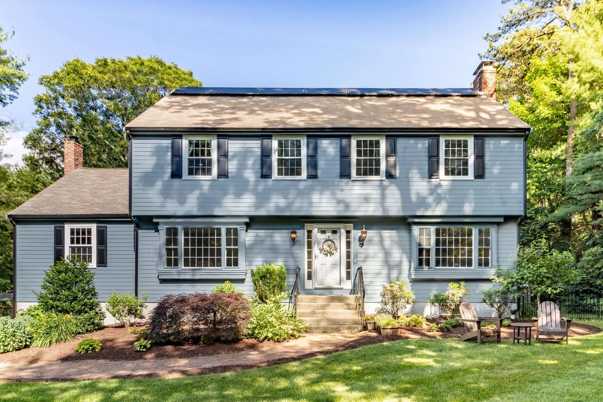 Two-story blue house with James Hardie siding, black shutters, central stairs, and two chairs on a sunny green lawn.