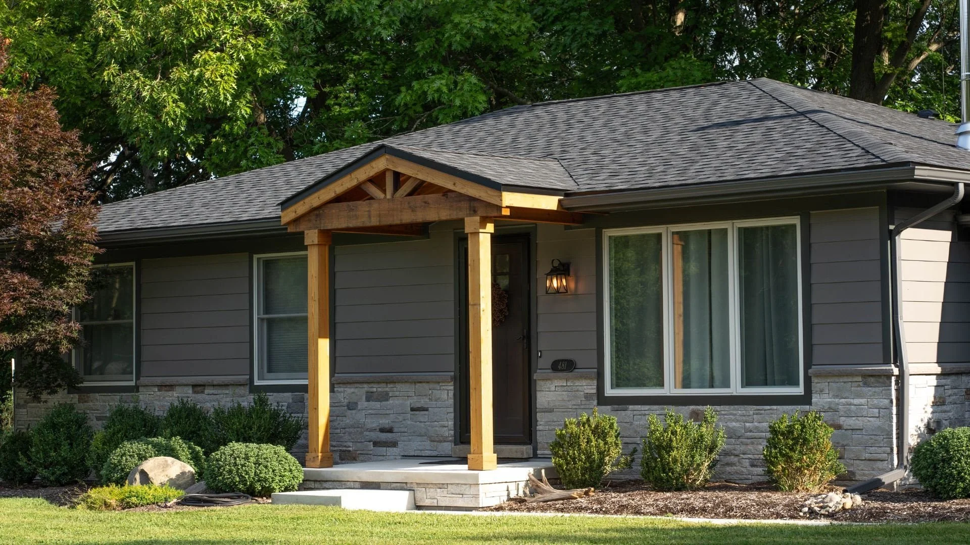 Single-story gray house with window replacement, wooden porch, stone accents, large windows, and a landscaped yard with green grass.