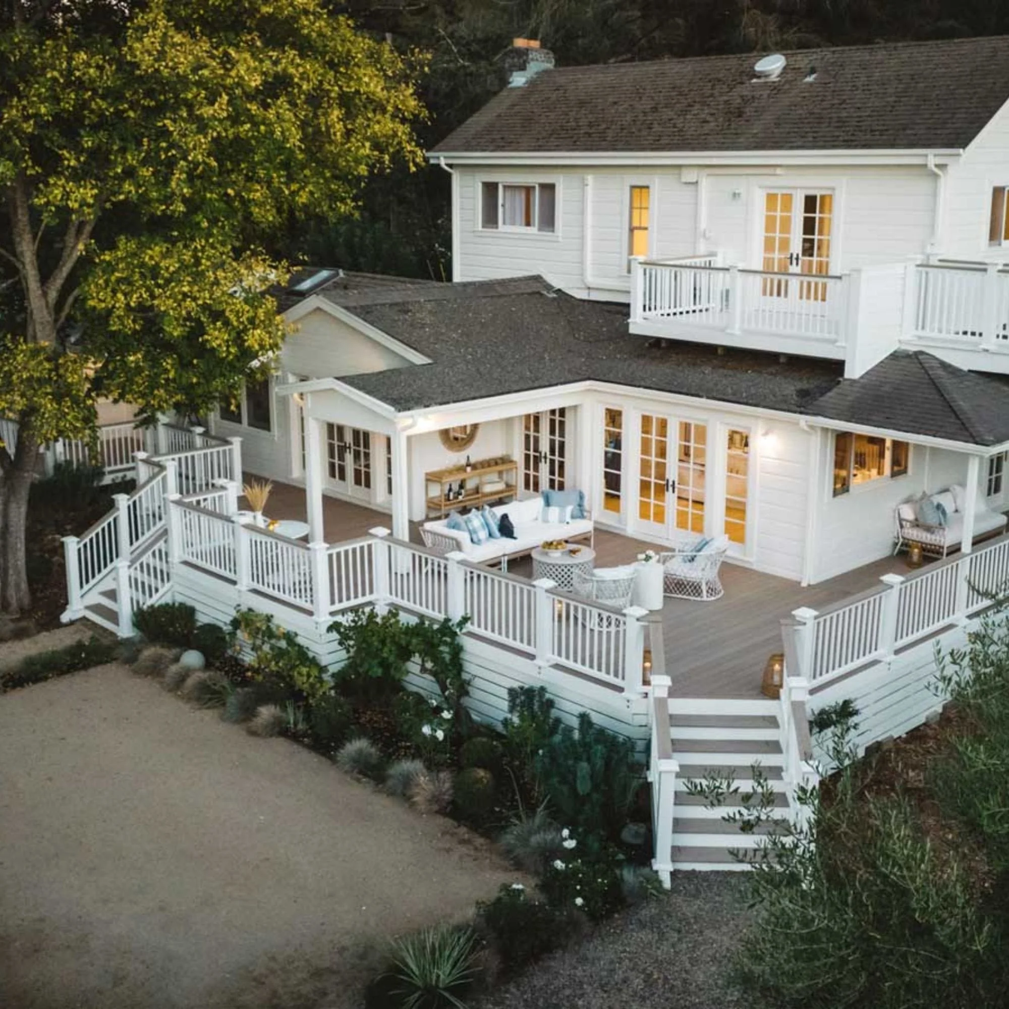 Aerial view of a white two-story house with a spacious wooden deck featuring outdoor seating, surrounded by a lush garden and trees.