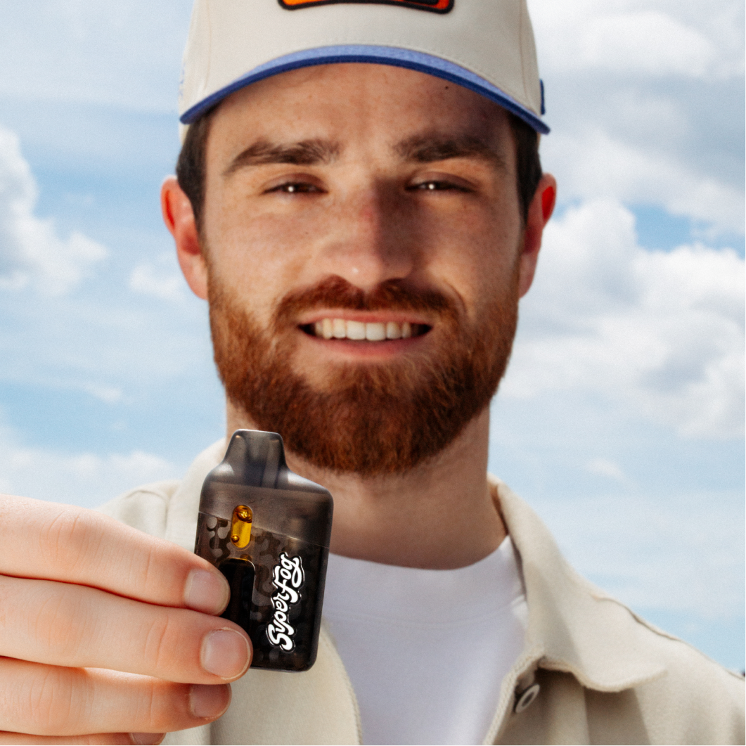 Man with a red beard holding a black vaporizer with 'Super Fog' written on it, smiling outdoors with blue sky and clouds in the background.