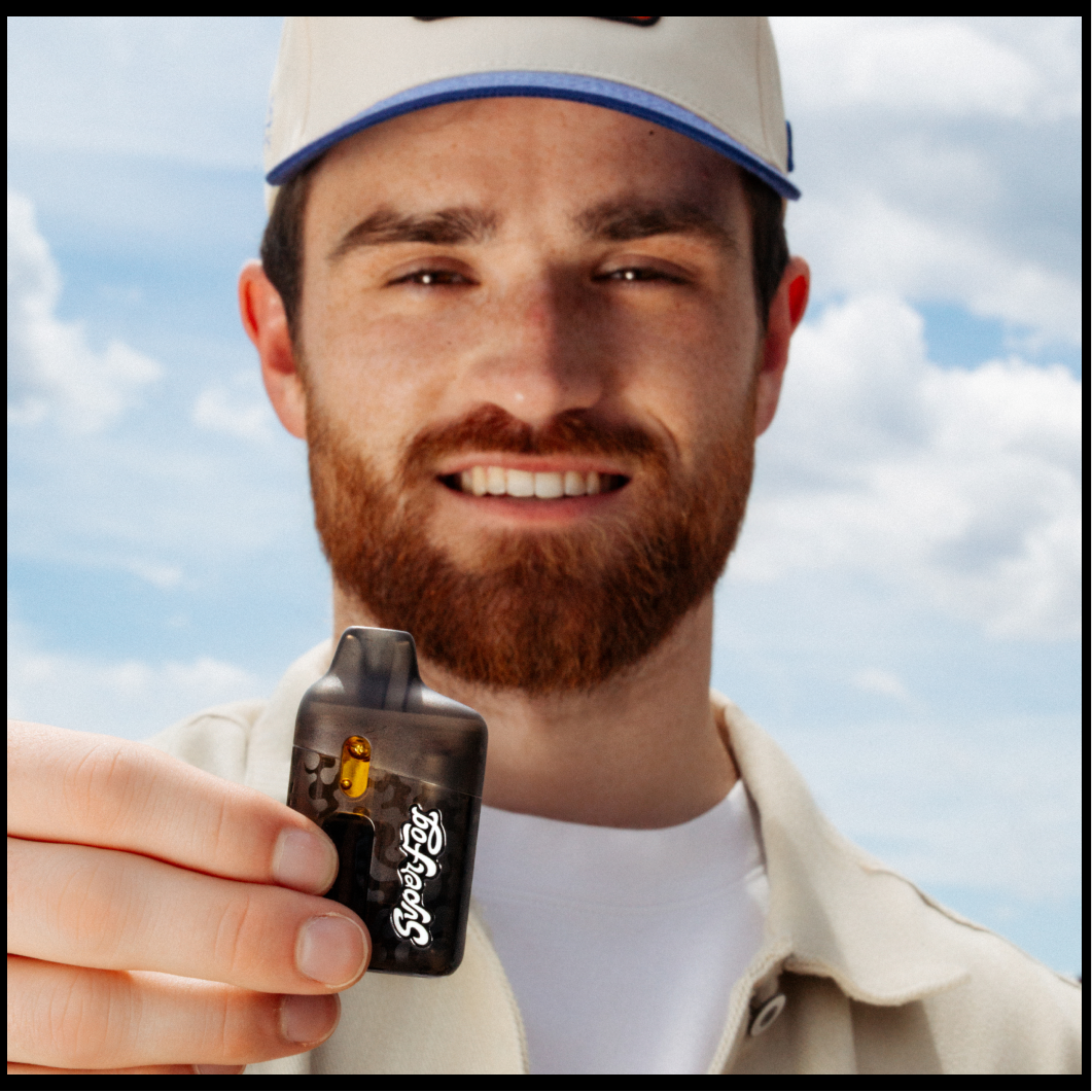 A man with a red beard and a baseball cap holding a small black lighter with the words 'Super Fog' written on it, outdoors with a partly cloudy sky in the background.