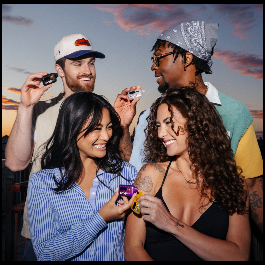 Four friends smiling and having fun during sunset, taking pictures with disposable cameras and one friend receiving a shot on her arm from another.
